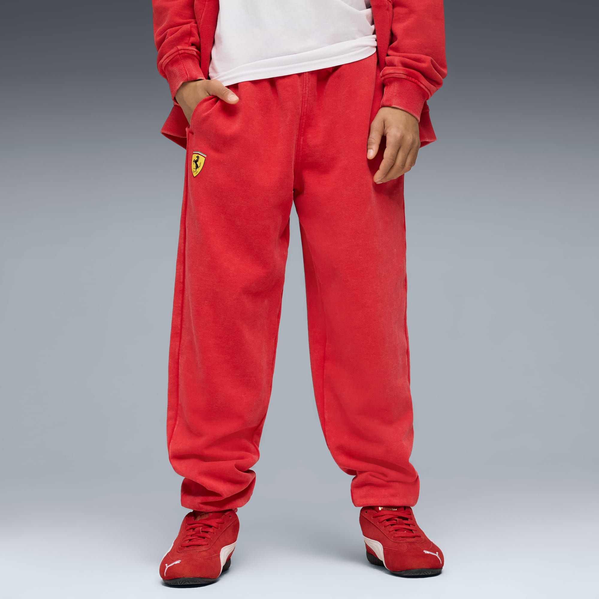 Tegen een grijze achtergrond draagt een model een rode broek, schoenen, jas en wit T-shirt.
