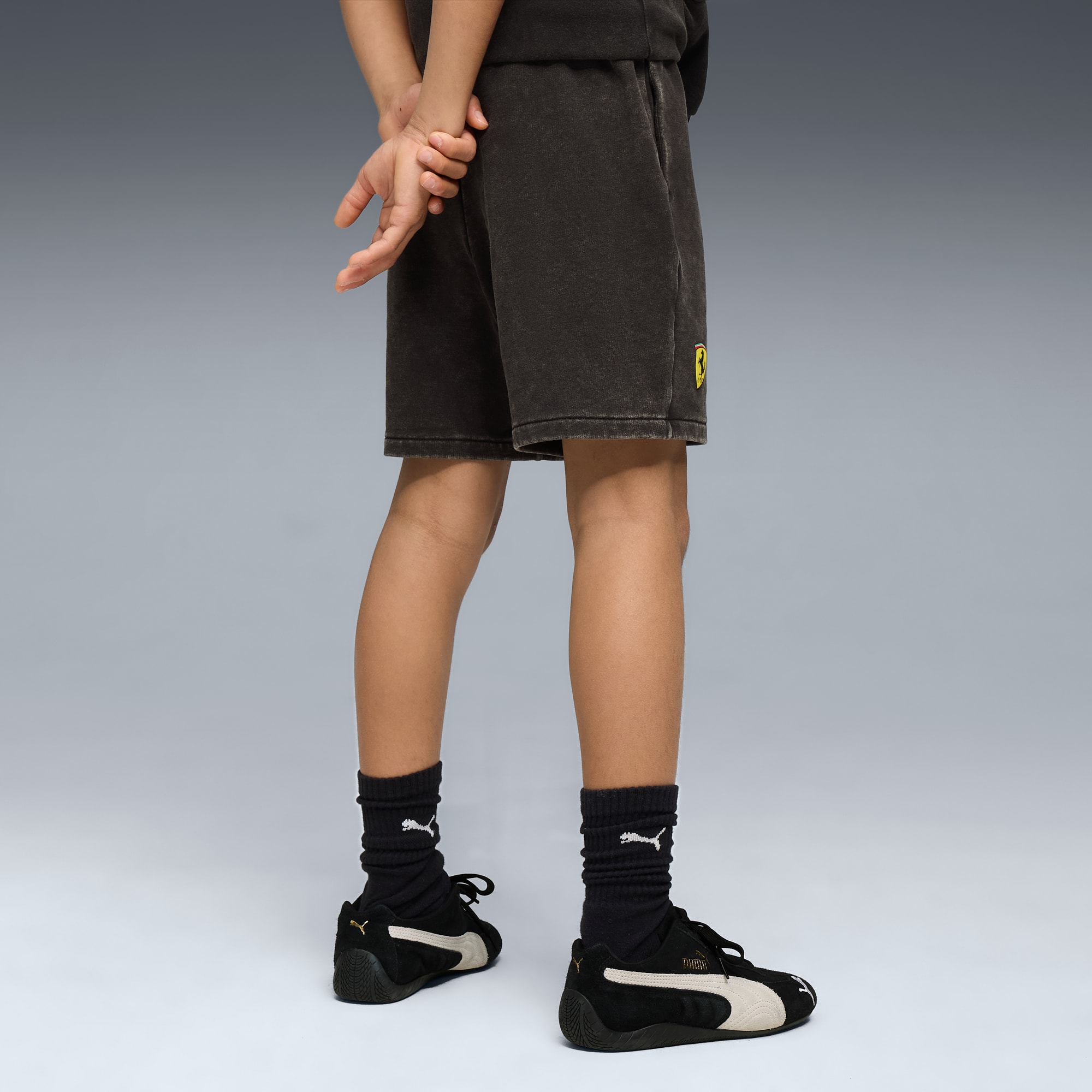 Na szarym tle dziecko prezentuje ciemne spodenki, skarpetki, tenisówki, ręce splecione za plecami.