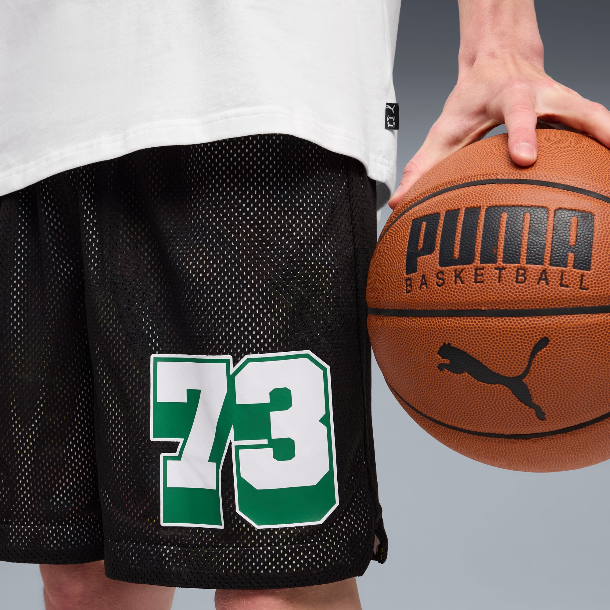 Su sfondo grigio, un modello tiene un pallone da basket, indossando una maglietta bianca e pantaloncini neri con il numero 73.