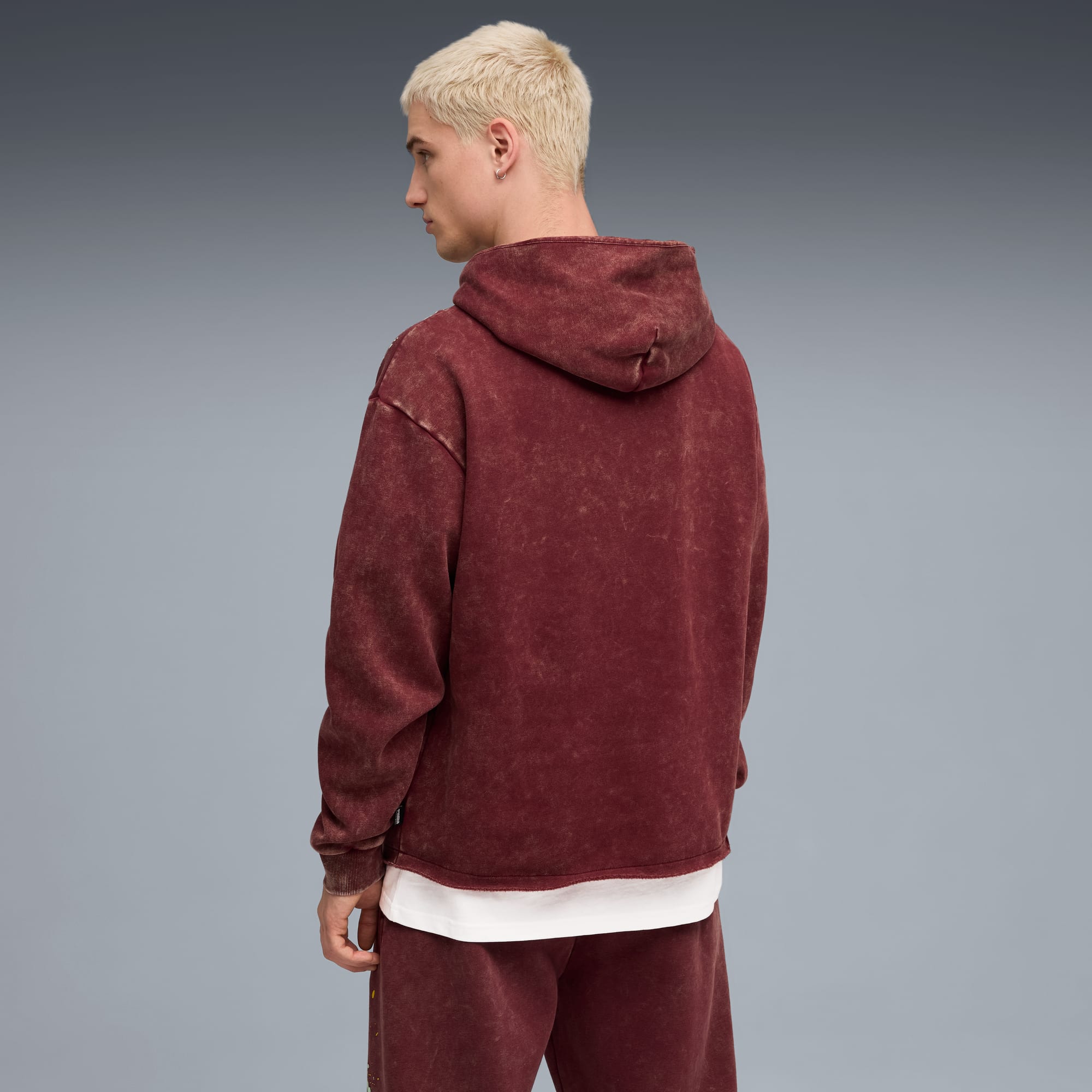 Sur fond gris, un modèle porte un sweat à capuche bordeaux et un pantalon assorti, regardant à gauche.