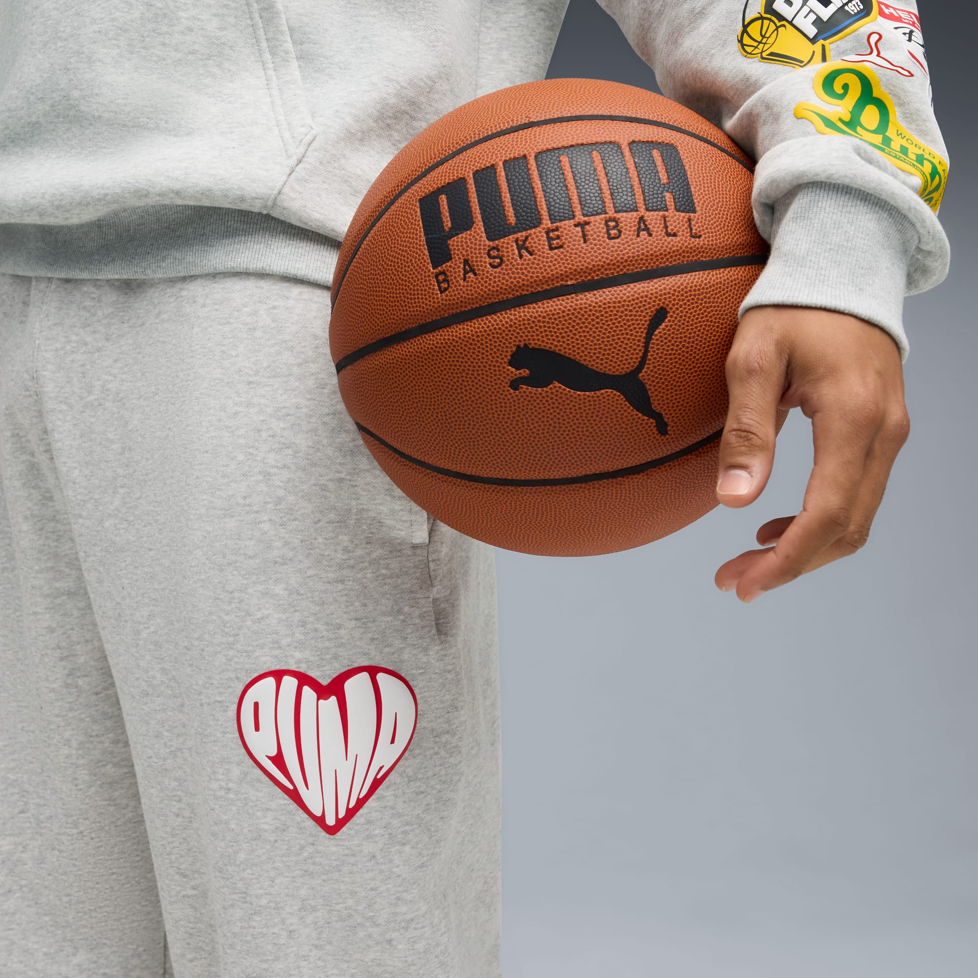 Na szarym tle osoba trzyma piłkę do koszykówki Puma, ubrana w szary dres.