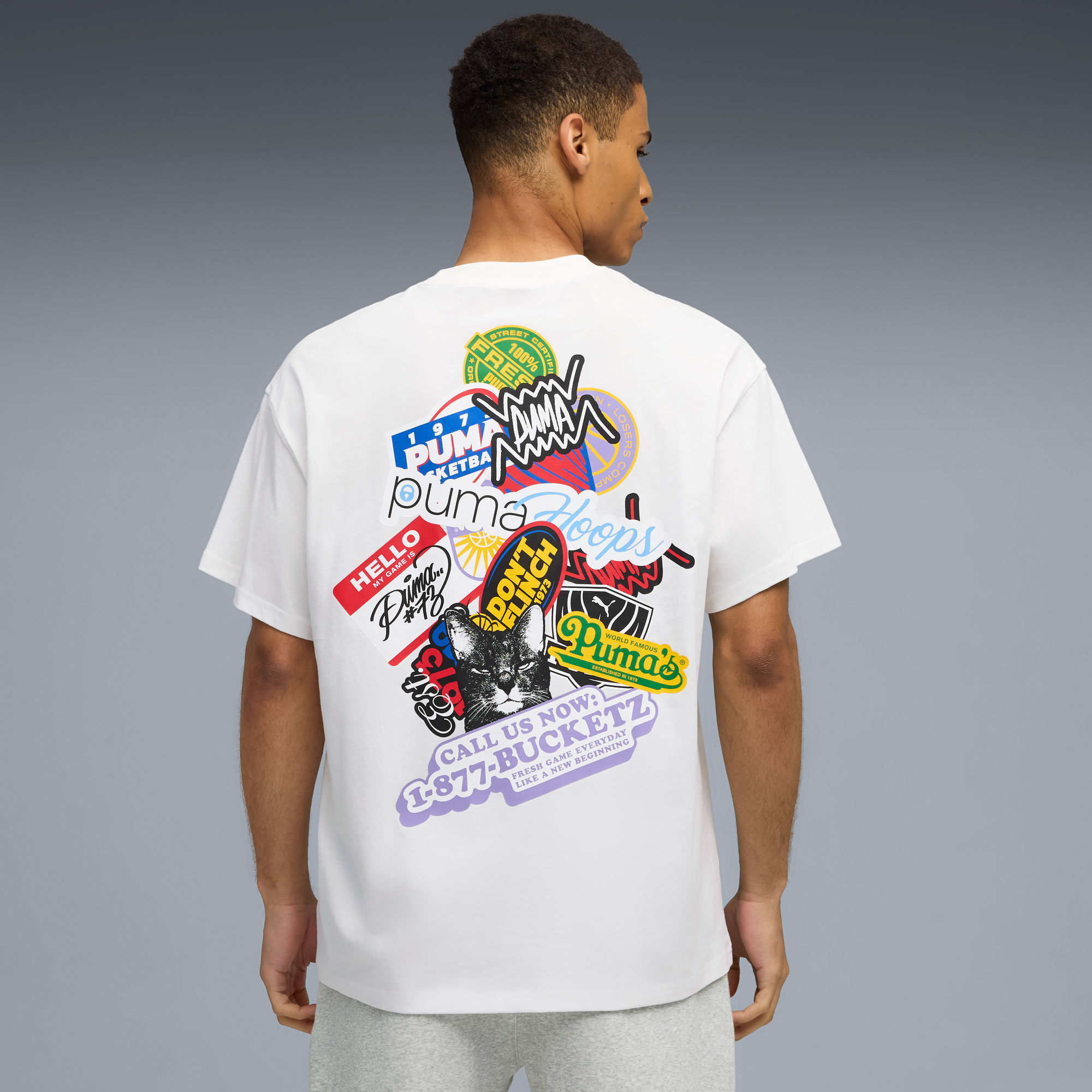 Een model poseert tegen een grijze achtergrond, gekleed in een wit t-shirt met sticker graphics.