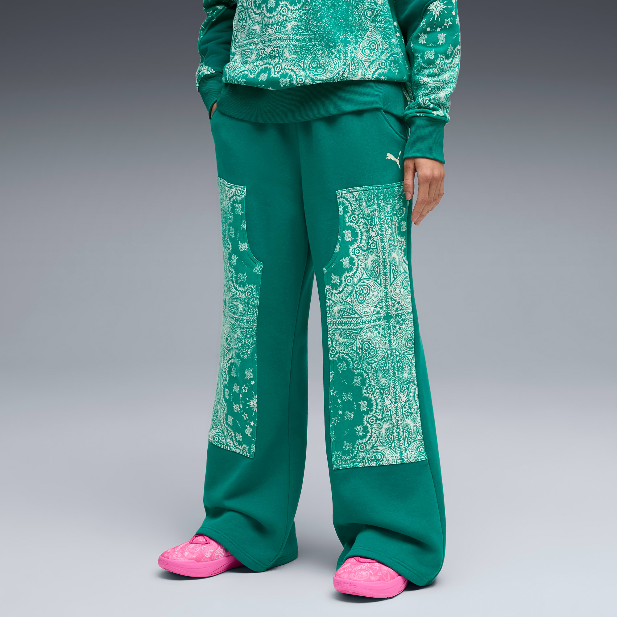 Vor grauem Hintergrund posiert ein Model in grüner Hose mit Paisleymuster und pinken Schuhen.