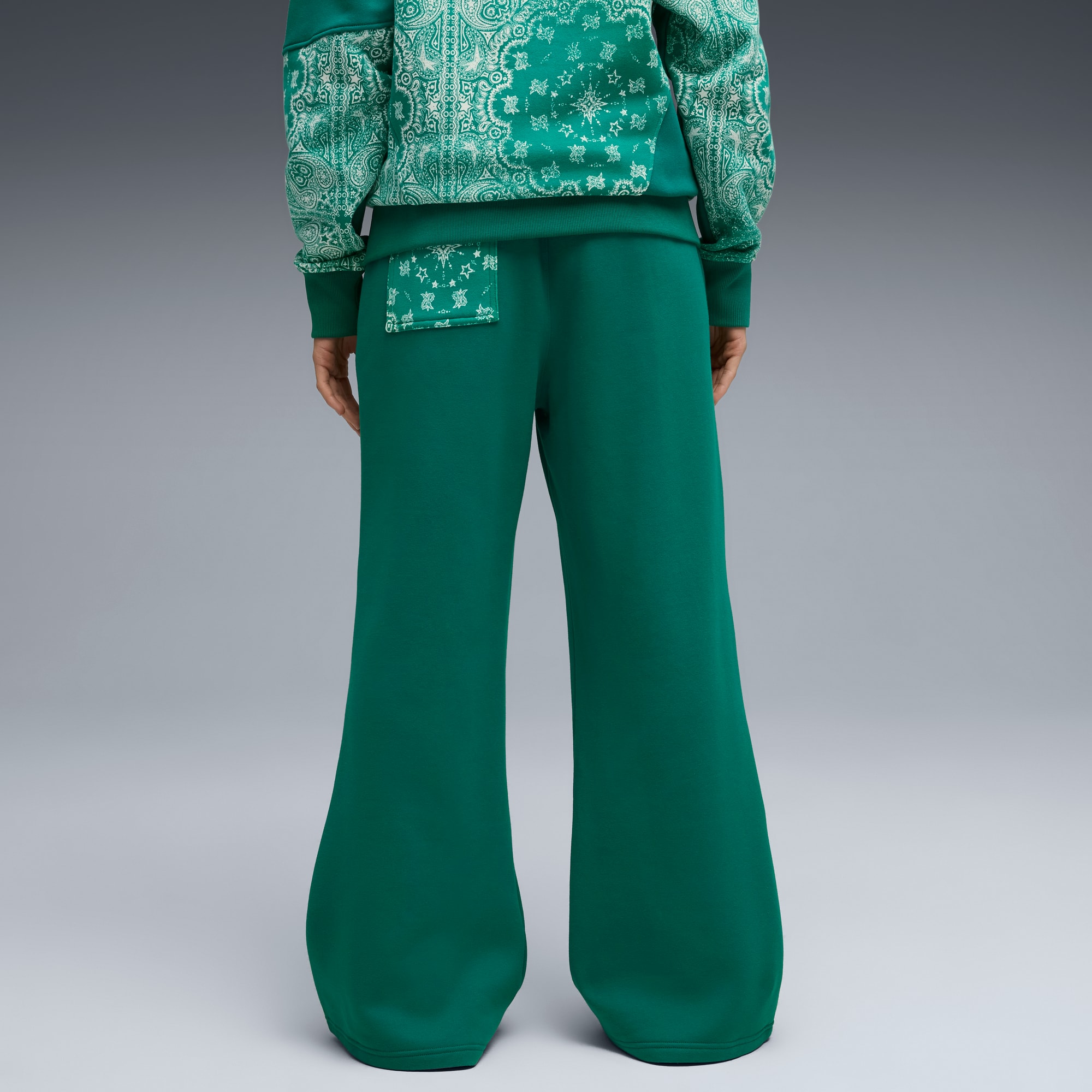 Ein Model steht vor einem grauen Hintergrund und trägt eine grüne Hose und ein gemustertes grünes Oberteil.