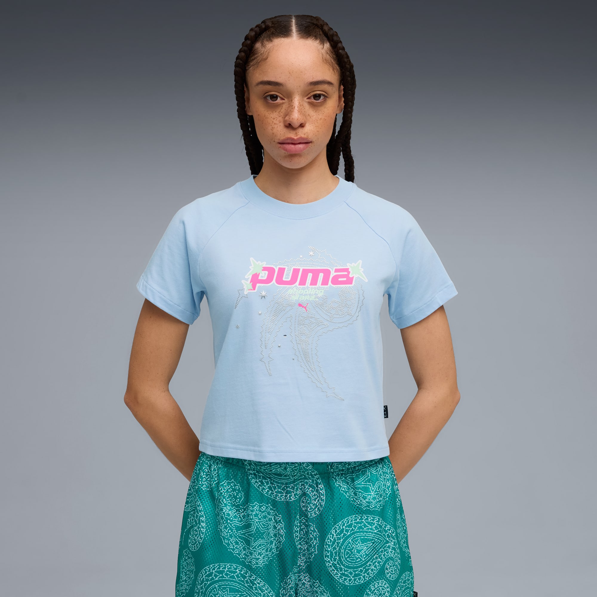 Sobre fondo gris, modelo posa con camiseta azul claro y pantalones cortos verdes estampados.