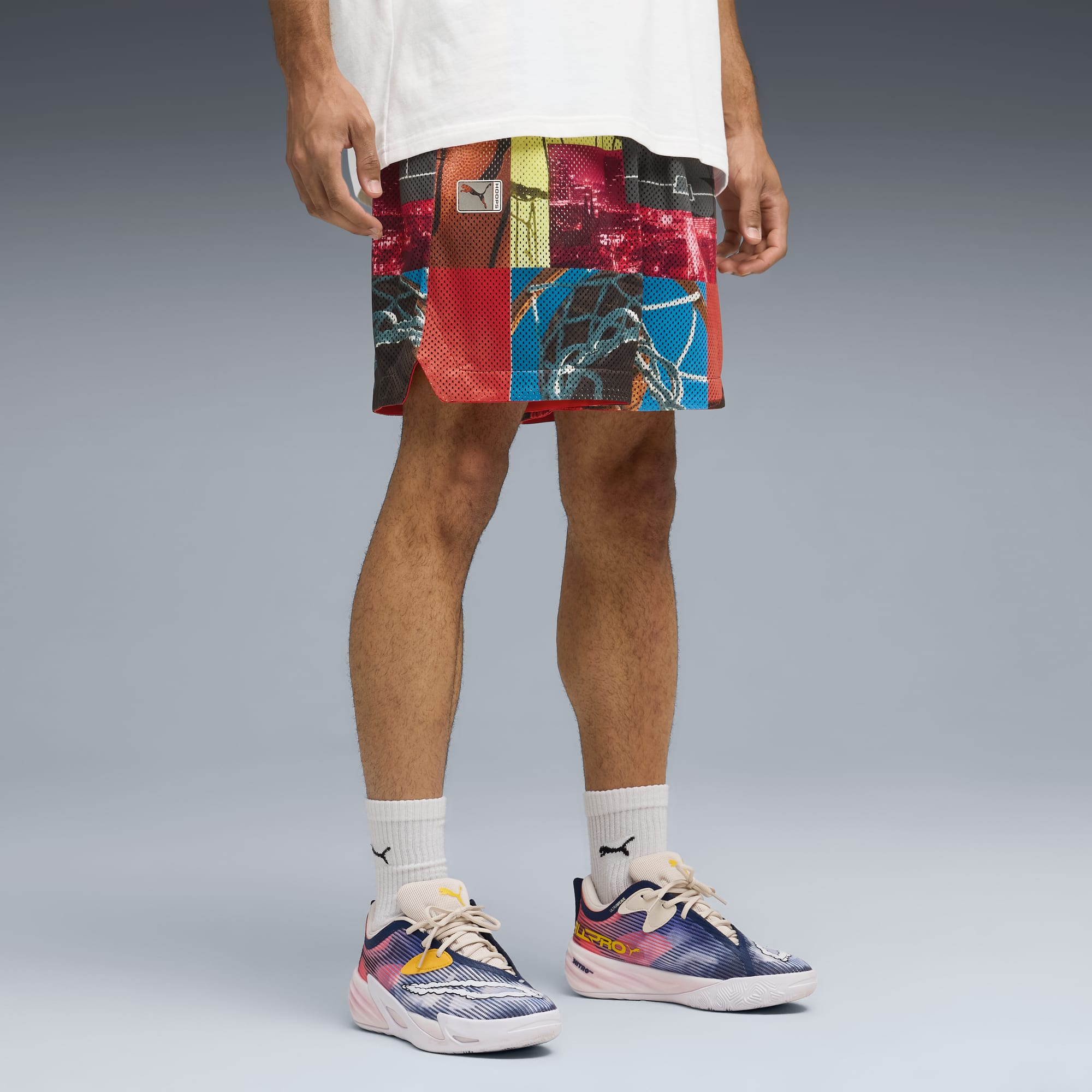 Vor grauem Hintergrund trägt ein Model ein weißes T-Shirt, gemusterte Shorts und Laufschuhe.