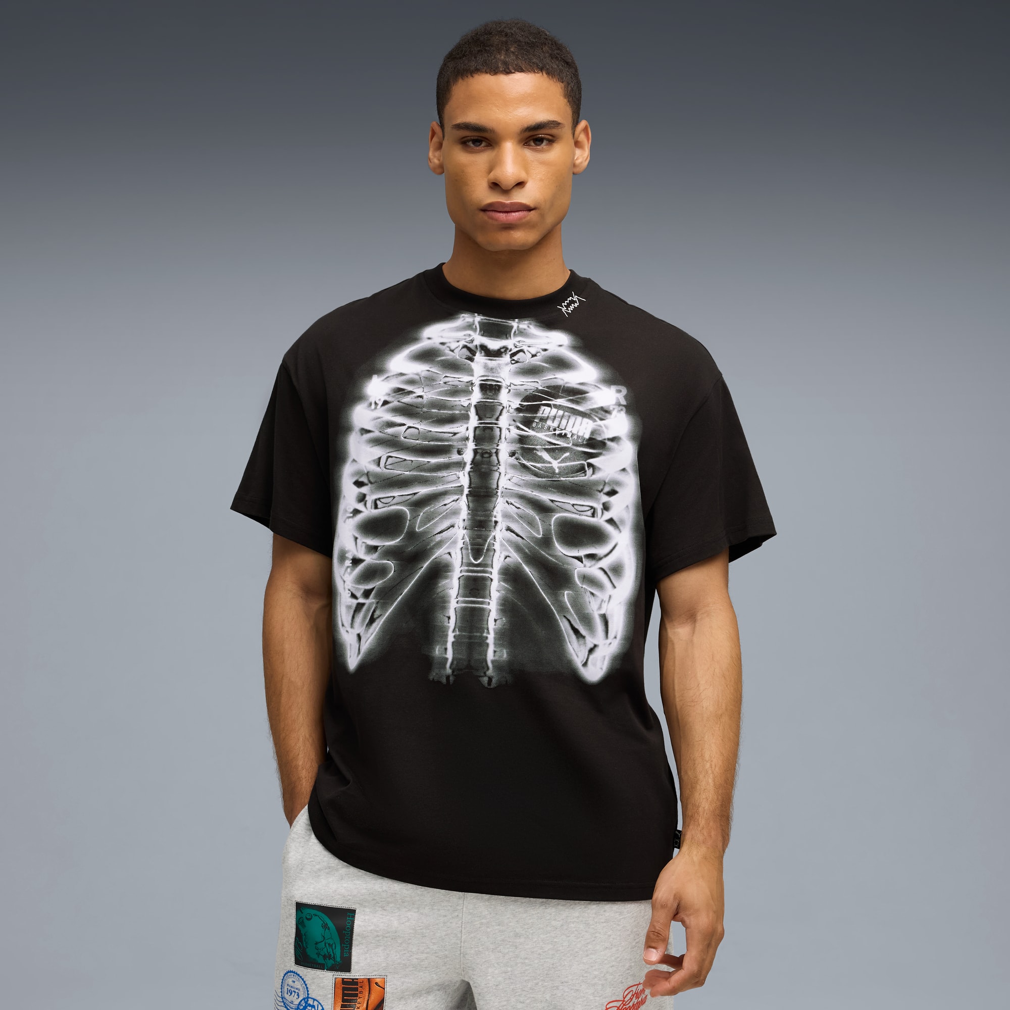 Vor grauem Hintergrund trägt ein Model ein schwarzes T-Shirt mit Skelett-Print und graue Hosen.