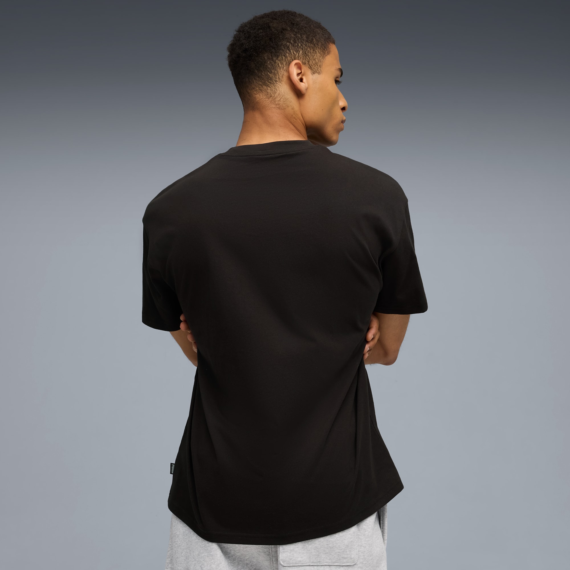 Ein Model mit verschränkten Armen blickt über die Schulter und trägt ein schwarzes T-Shirt und graue Shorts.