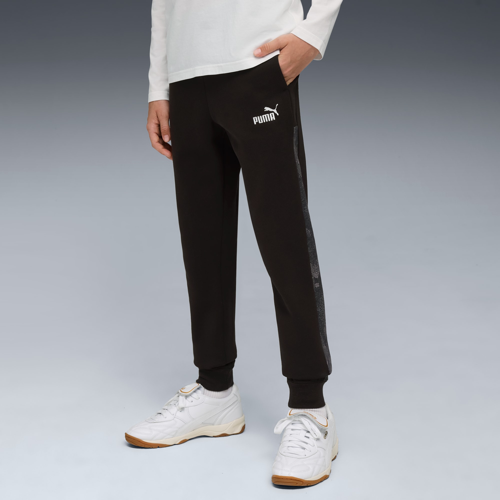 Een model poseert tegen grijs met een zwarte broek, witte top en witte sportschoenen.