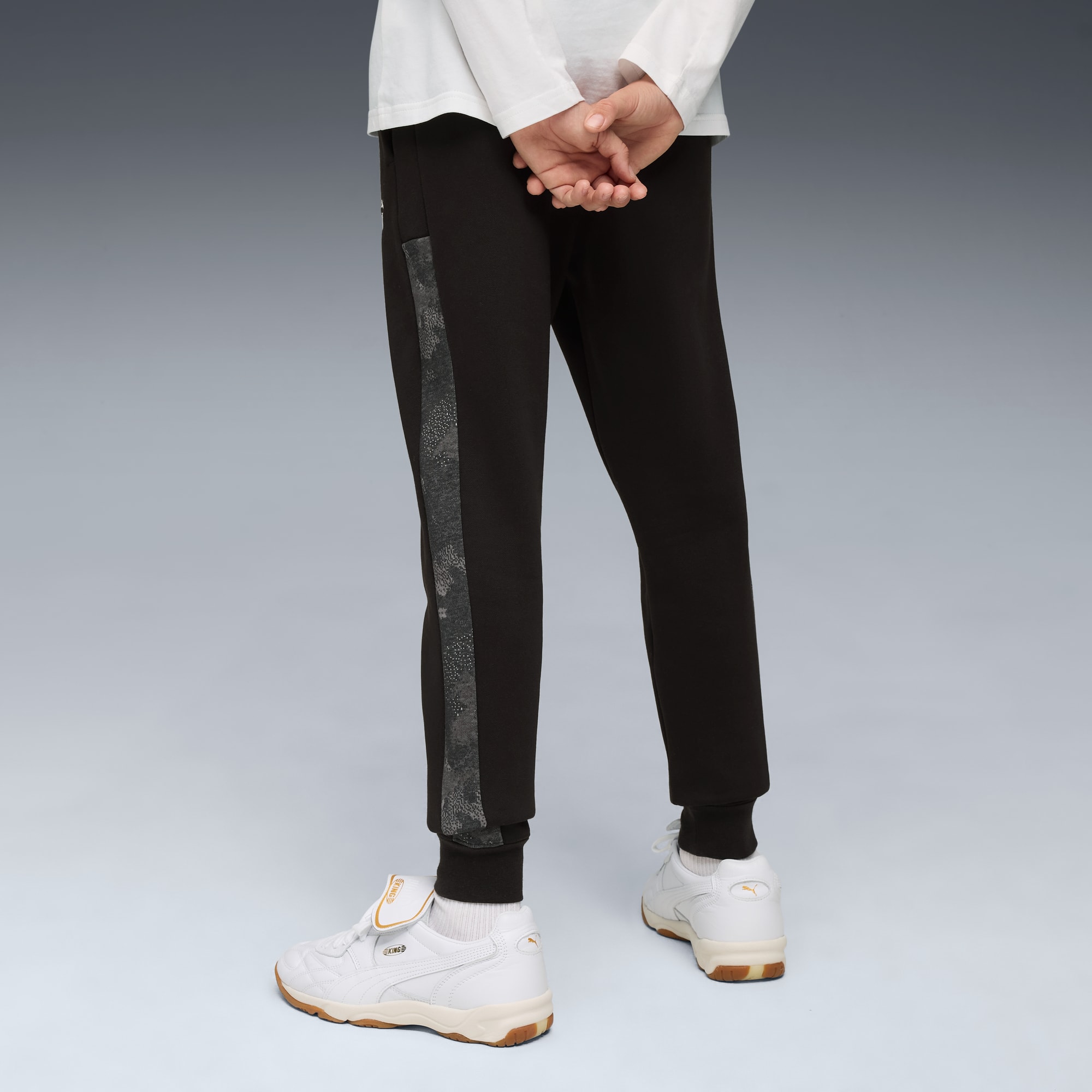 Tegen een grijze achtergrond draagt een model een zwarte joggingbroek met een zijstreep en witte sportschoenen.