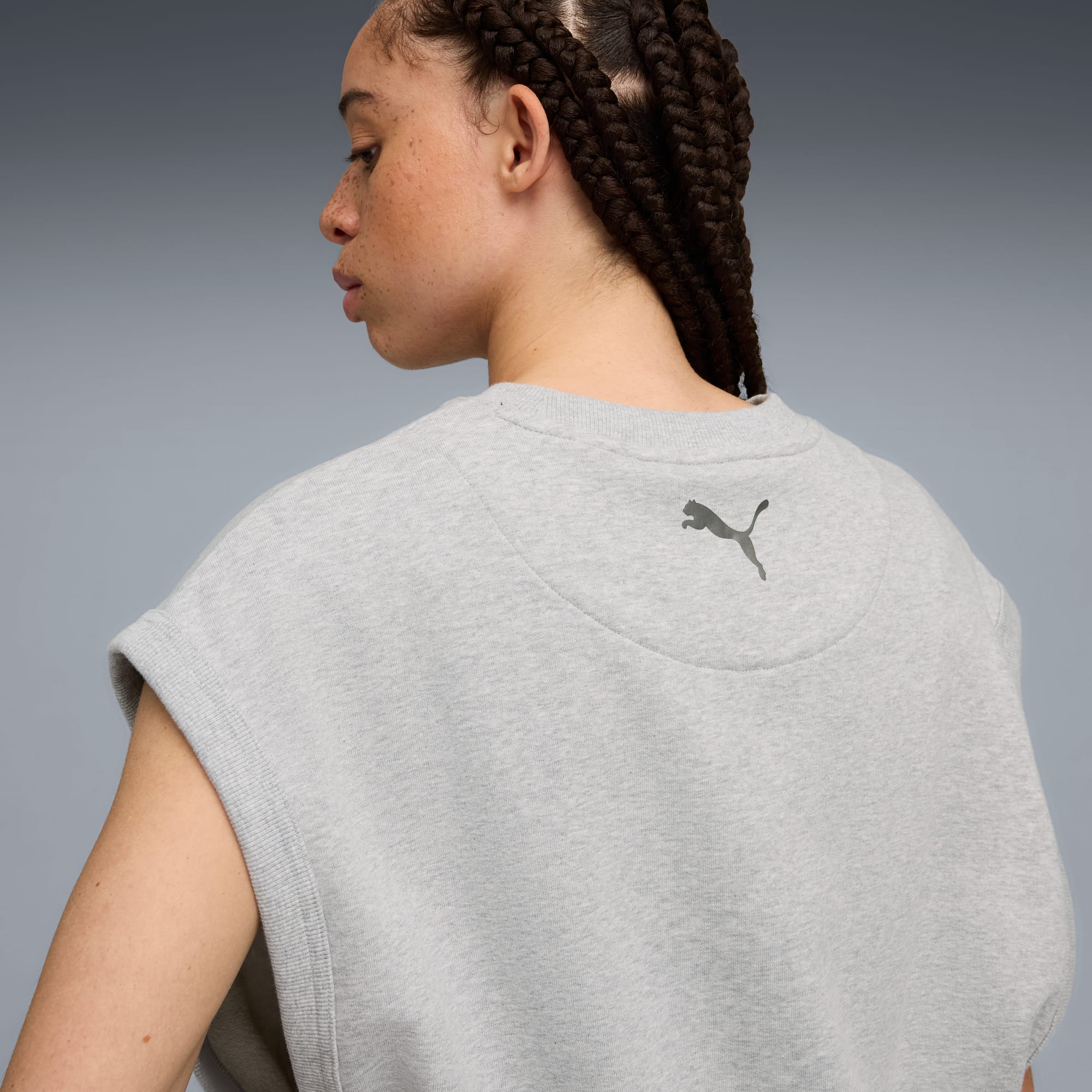 Een model met gevlochten haar poseert tegen een grijze achtergrond, gekleed in een grijze mouwloze top.