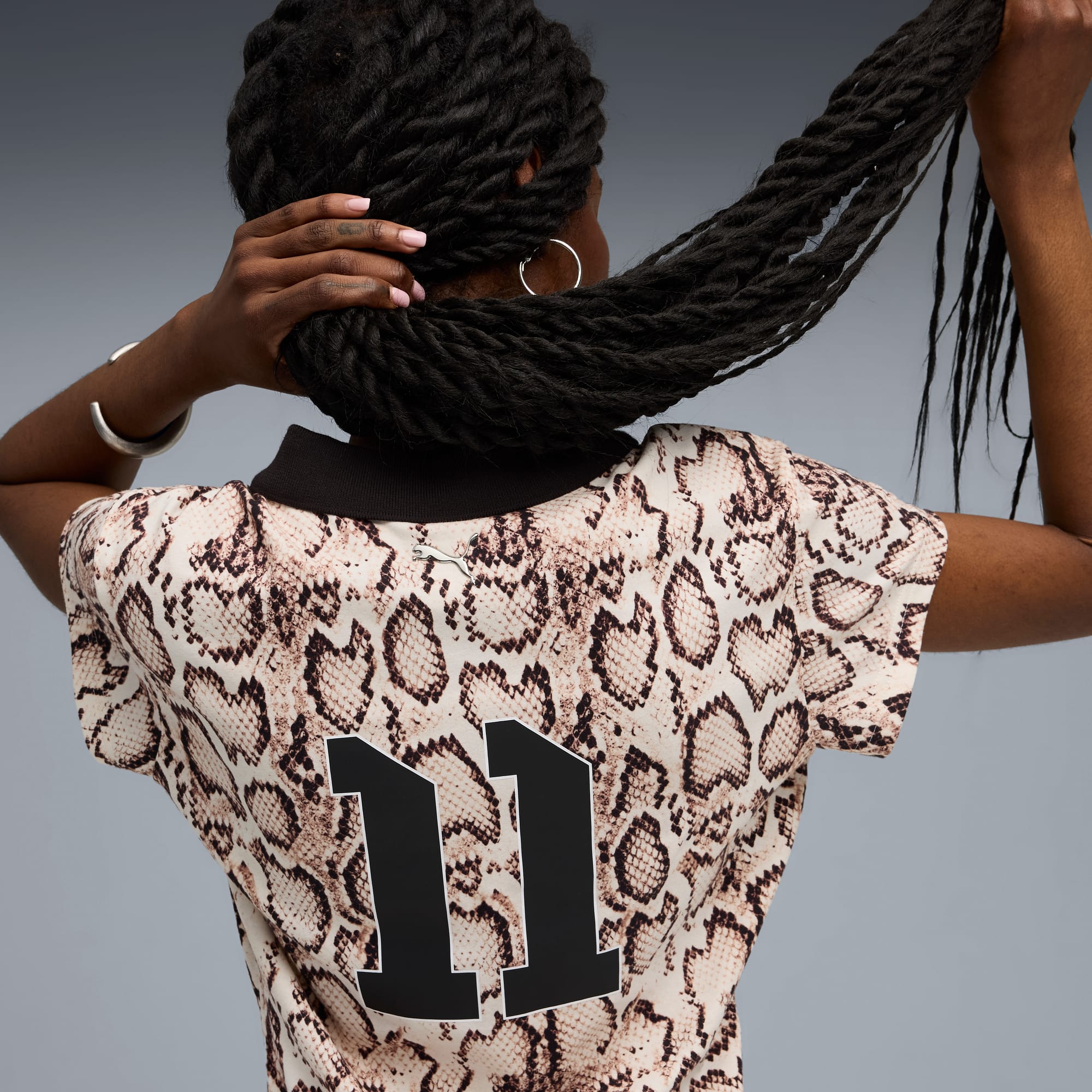 Tegen een grijze achtergrond raakt een model haar haar aan, ze draagt een top met slangenprint en nummer 11.