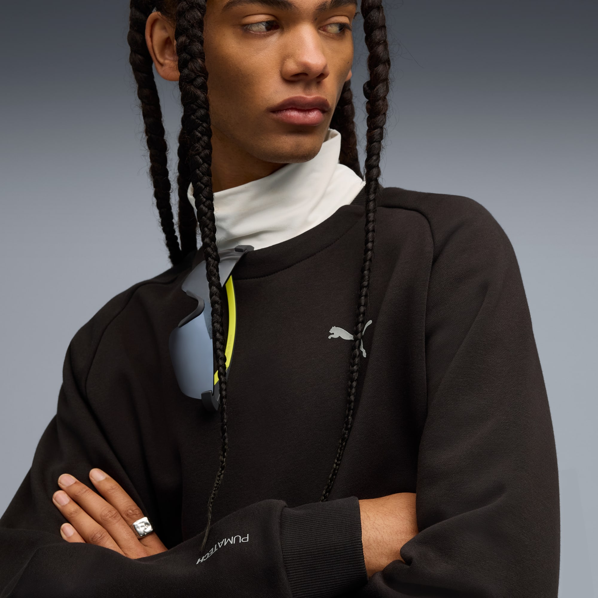 Na szarym tle model z warkoczami ma na sobie czarną bluzę i okulary przeciwsłoneczne.