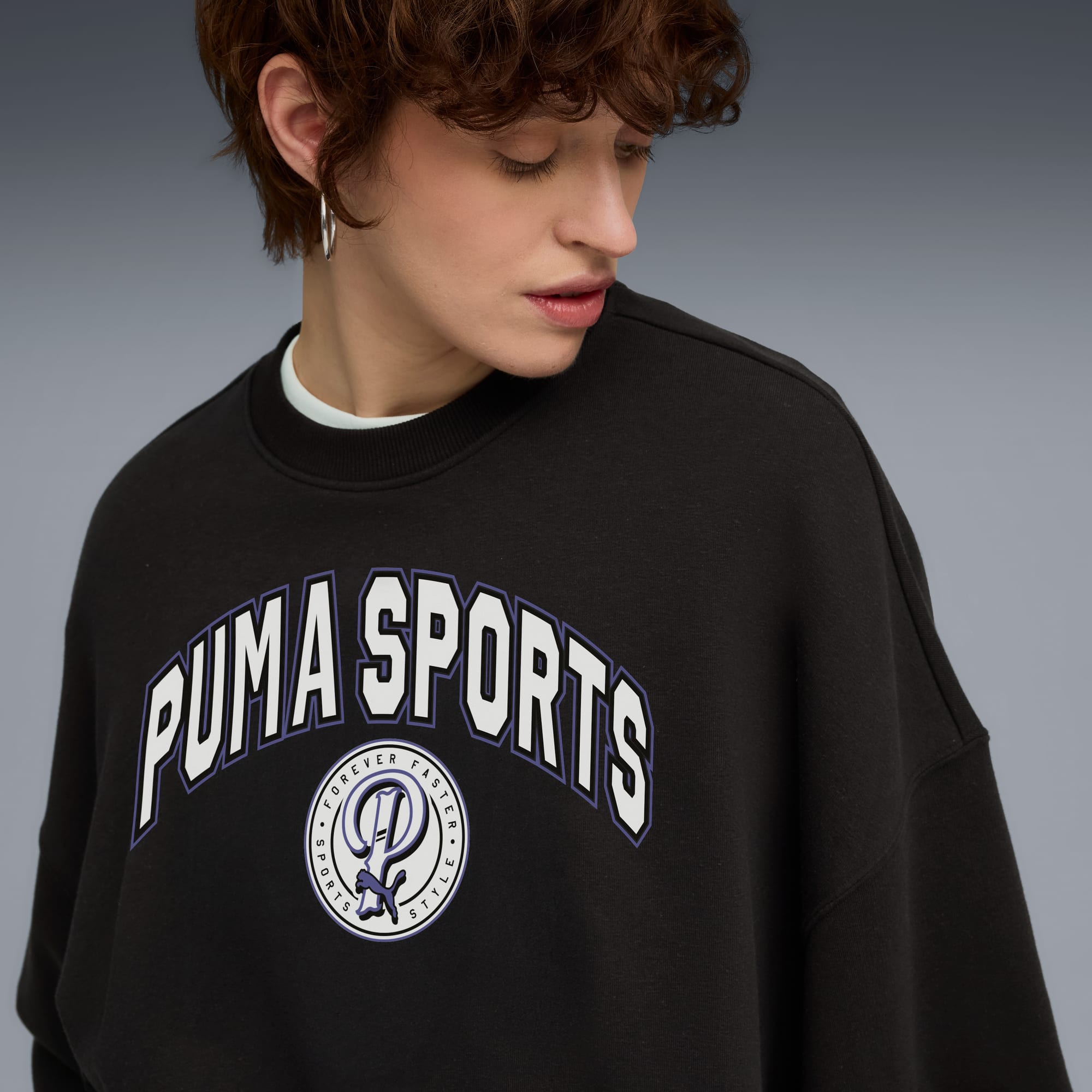 Model voor een grijze achtergrond met een zwart sweatshirt met 'PUMA SPORTS'-tekst.