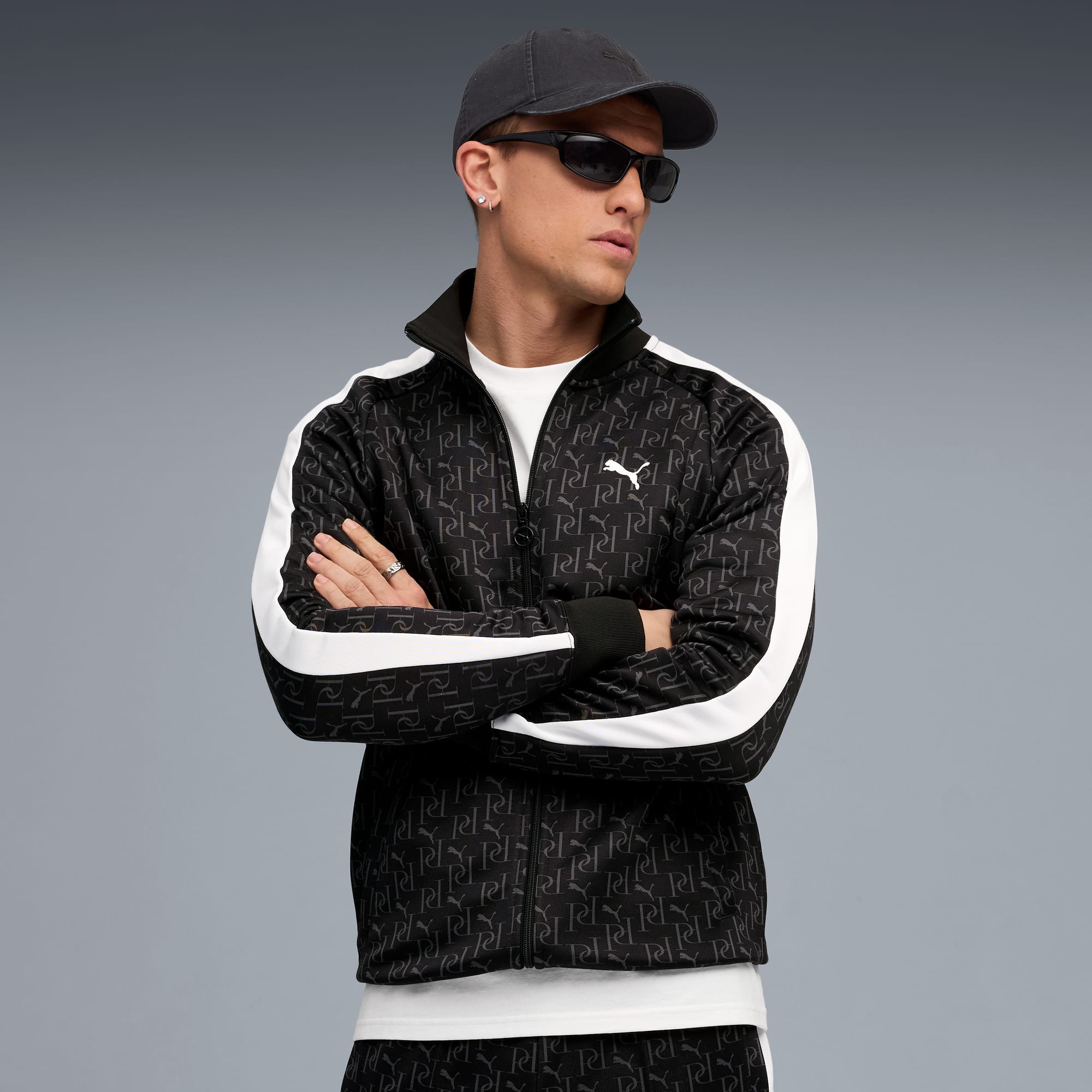 Un mannequin, bras croisés, porte veste à motifs, casquette et lunettes sur fond gris.
