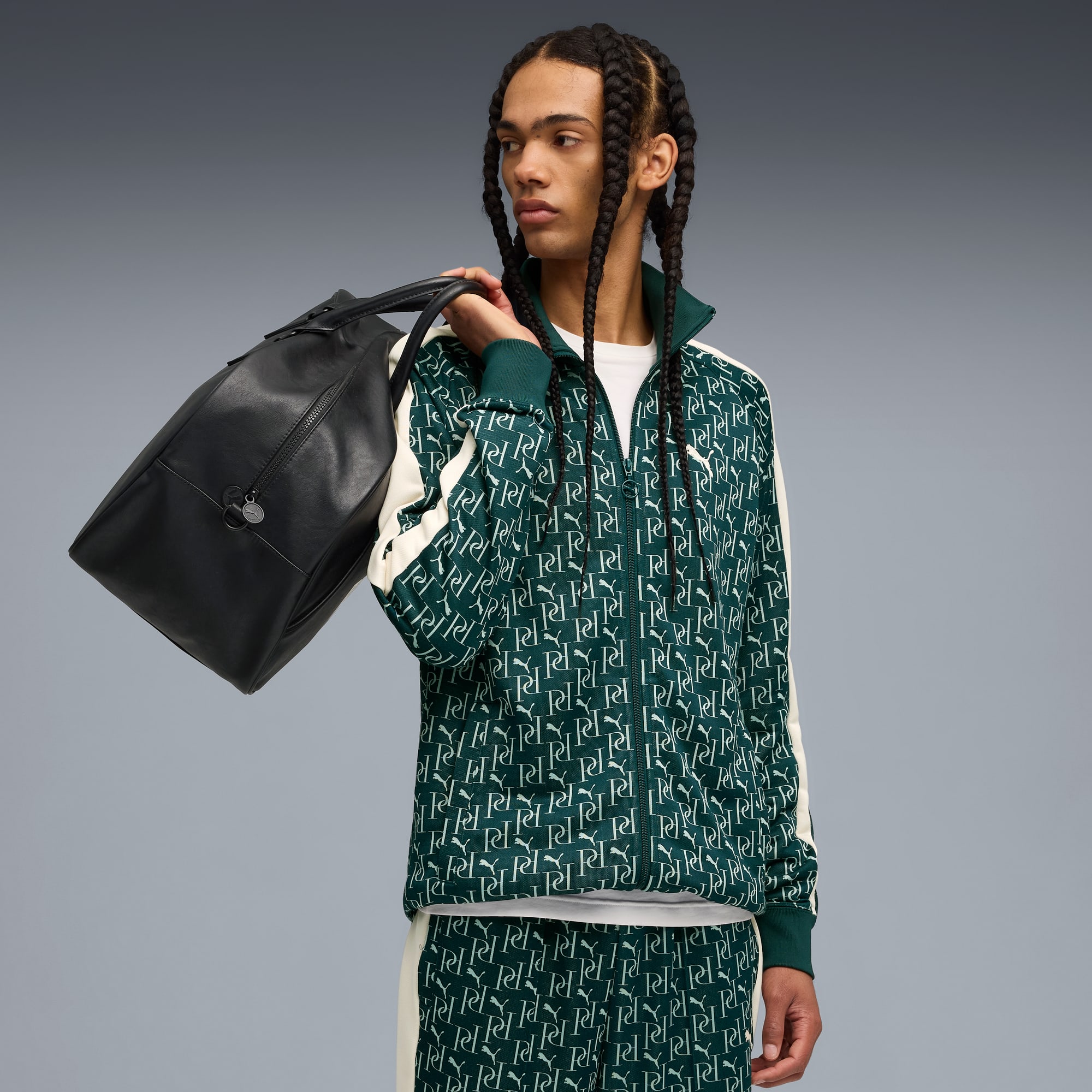 Sur fond gris, un mannequin en survêtement vert à motifs porte un sac noir.