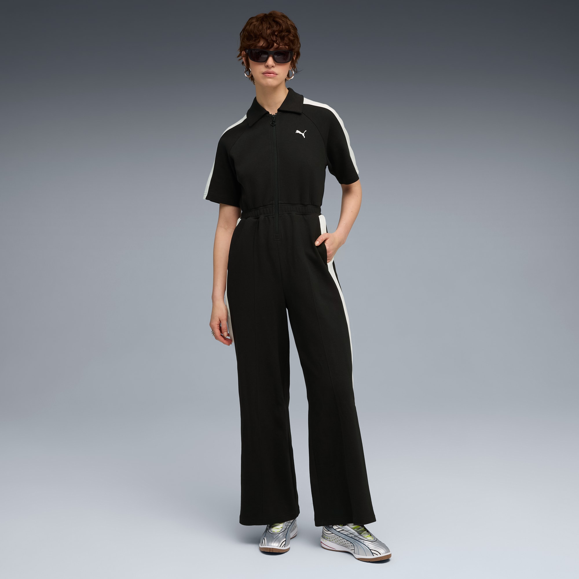Een model staat op een grijze achtergrond in een zwarte jumpsuit met witte strepen.