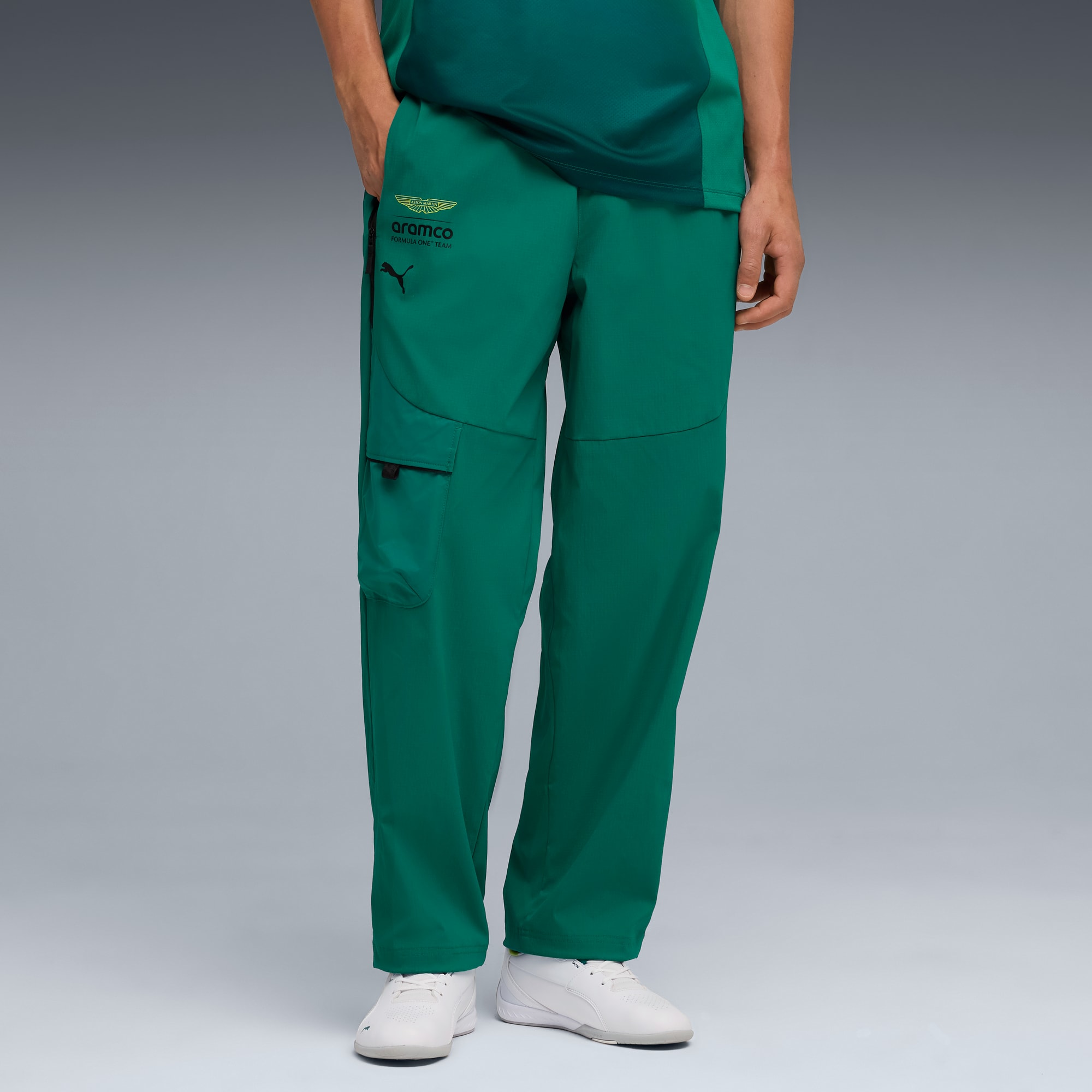 Vor grauem Hintergrund posiert ein Model mit der Hand in der Tasche in grüner Hose und weißen Turnschuhen.