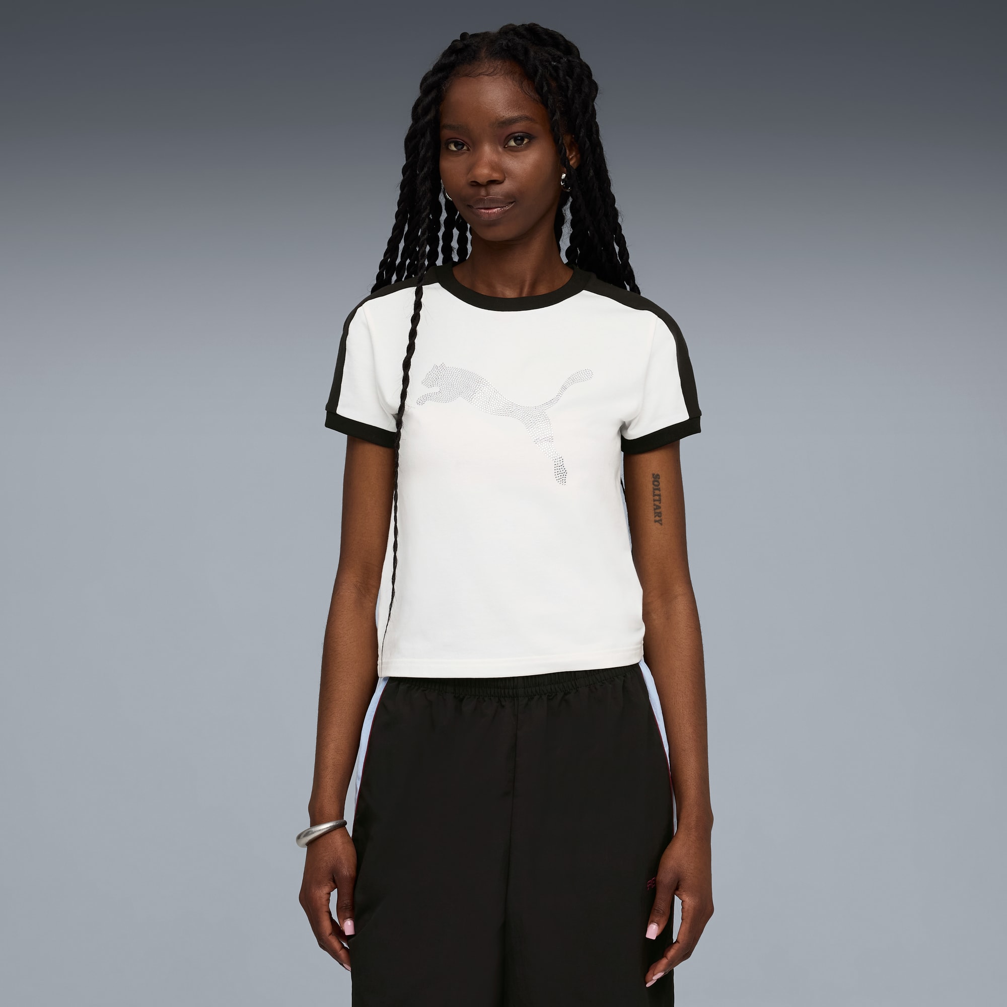 Una modella posa su uno sfondo grigio, indossando un top bianco e pantaloni neri.