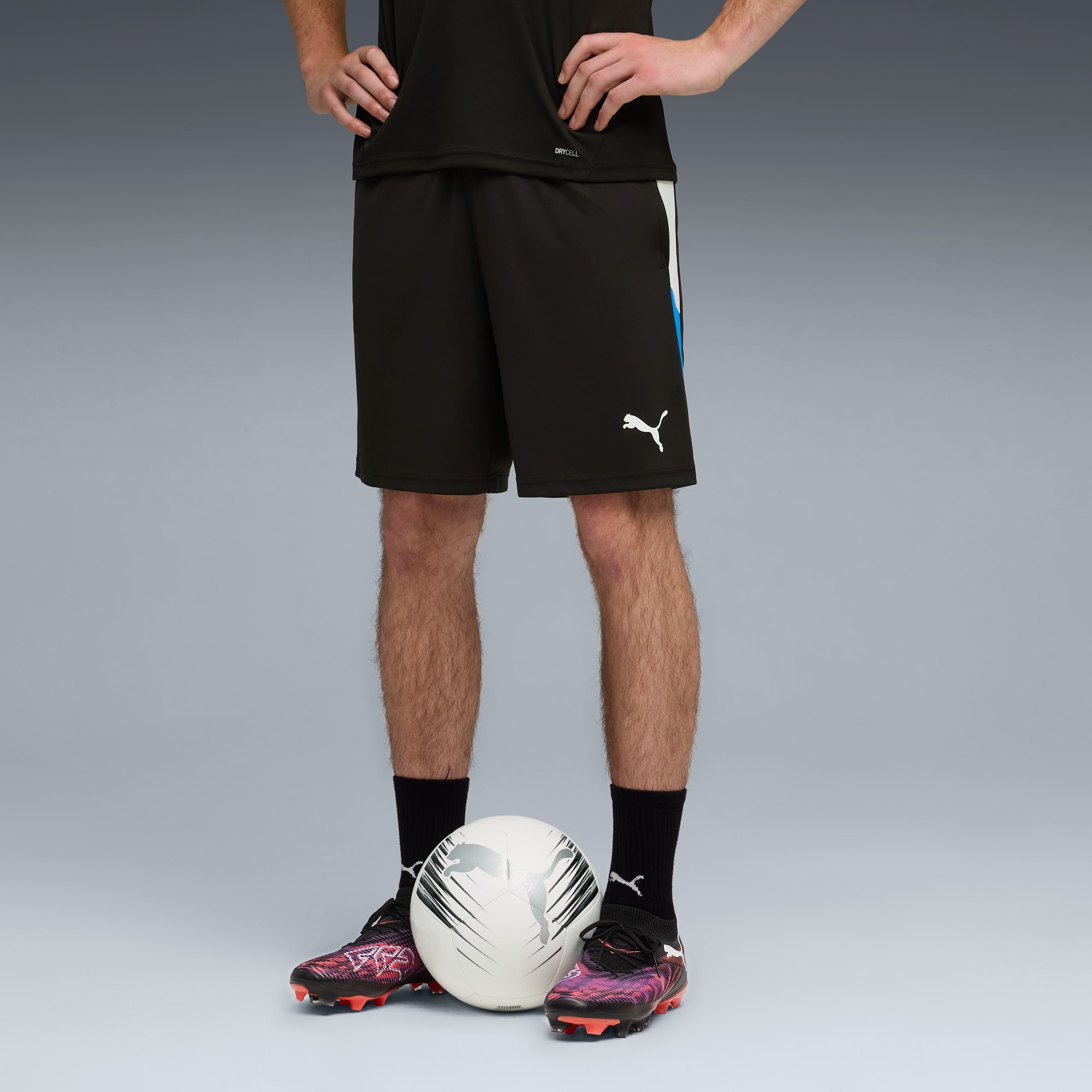 Ein Model steht mit einem Fußball, trägt schwarze Shorts, Socken und bunte Fußballschuhe.