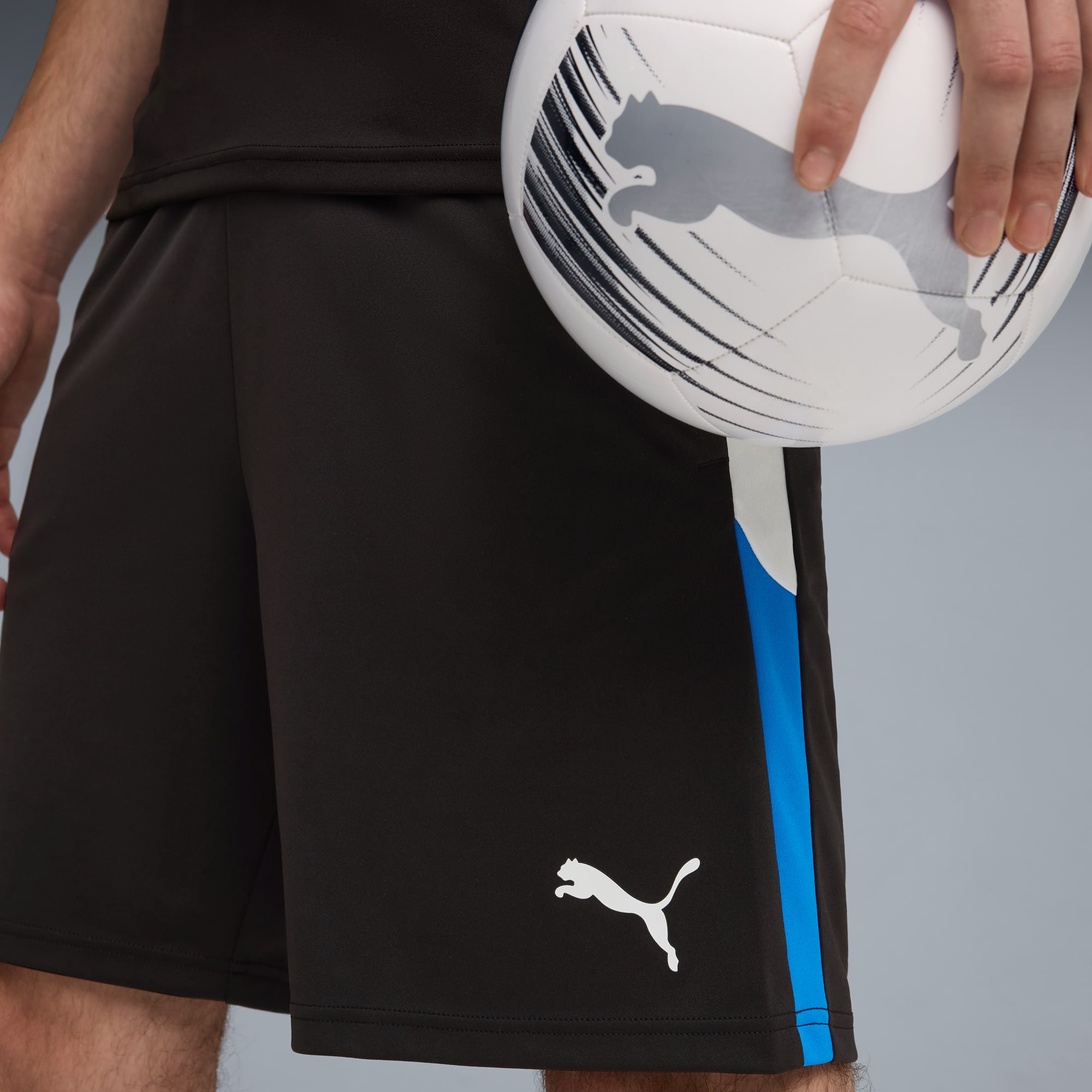 Vor grauem Hintergrund hält ein Model einen Puma-Ball und trägt schwarze Shorts mit blauen Details.