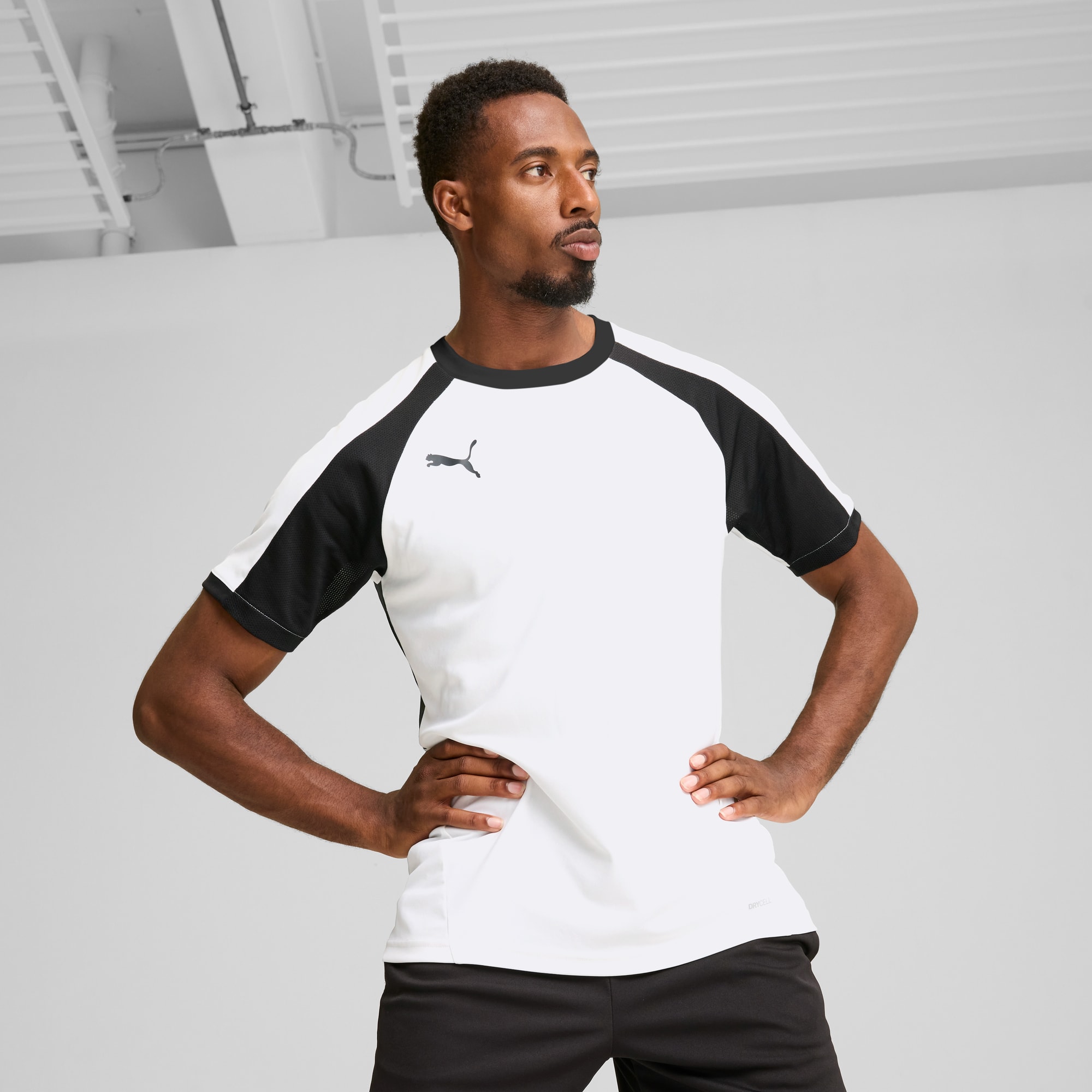 Ein Model posiert vor grauem Hintergrund in einem weiss-schwarzen Puma T-Shirt und schwarzen Shorts.