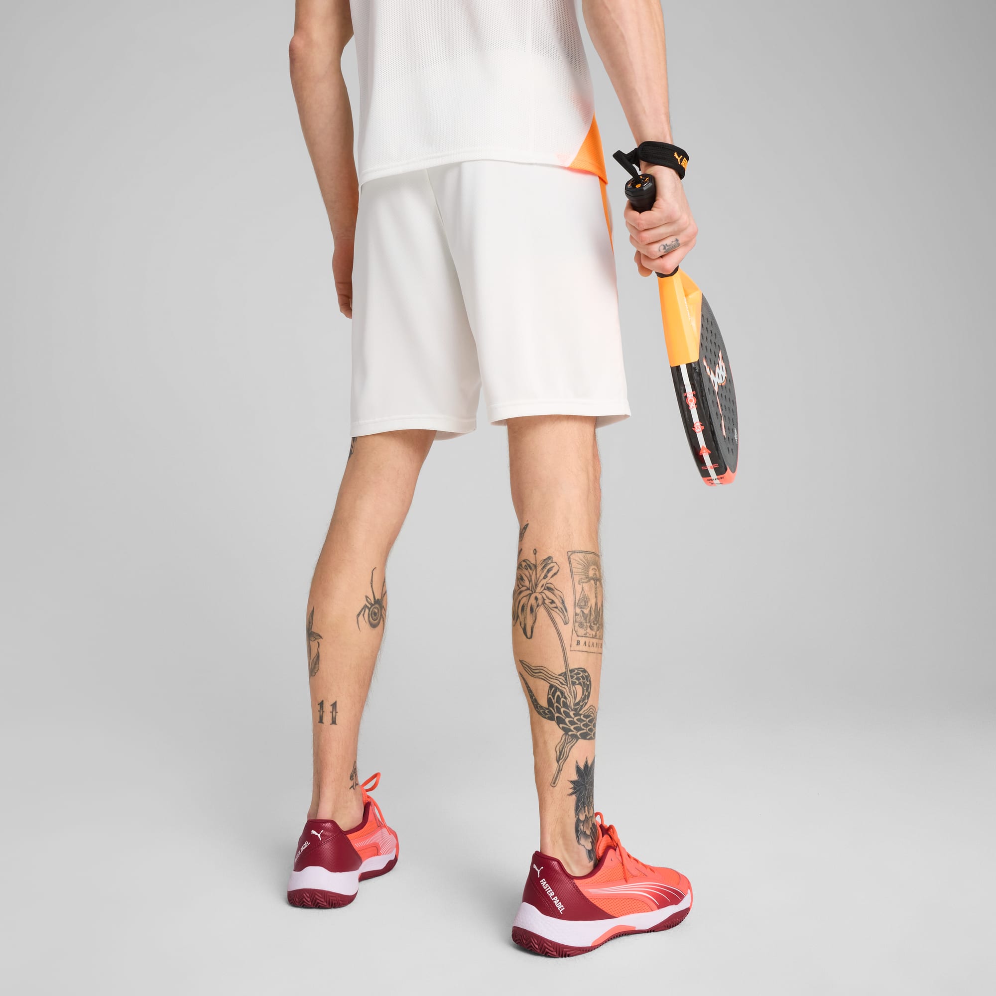 Ein Model vor grau hält einen Schläger; trägt ein weißes Top, Shorts und rote Turnschuhe.