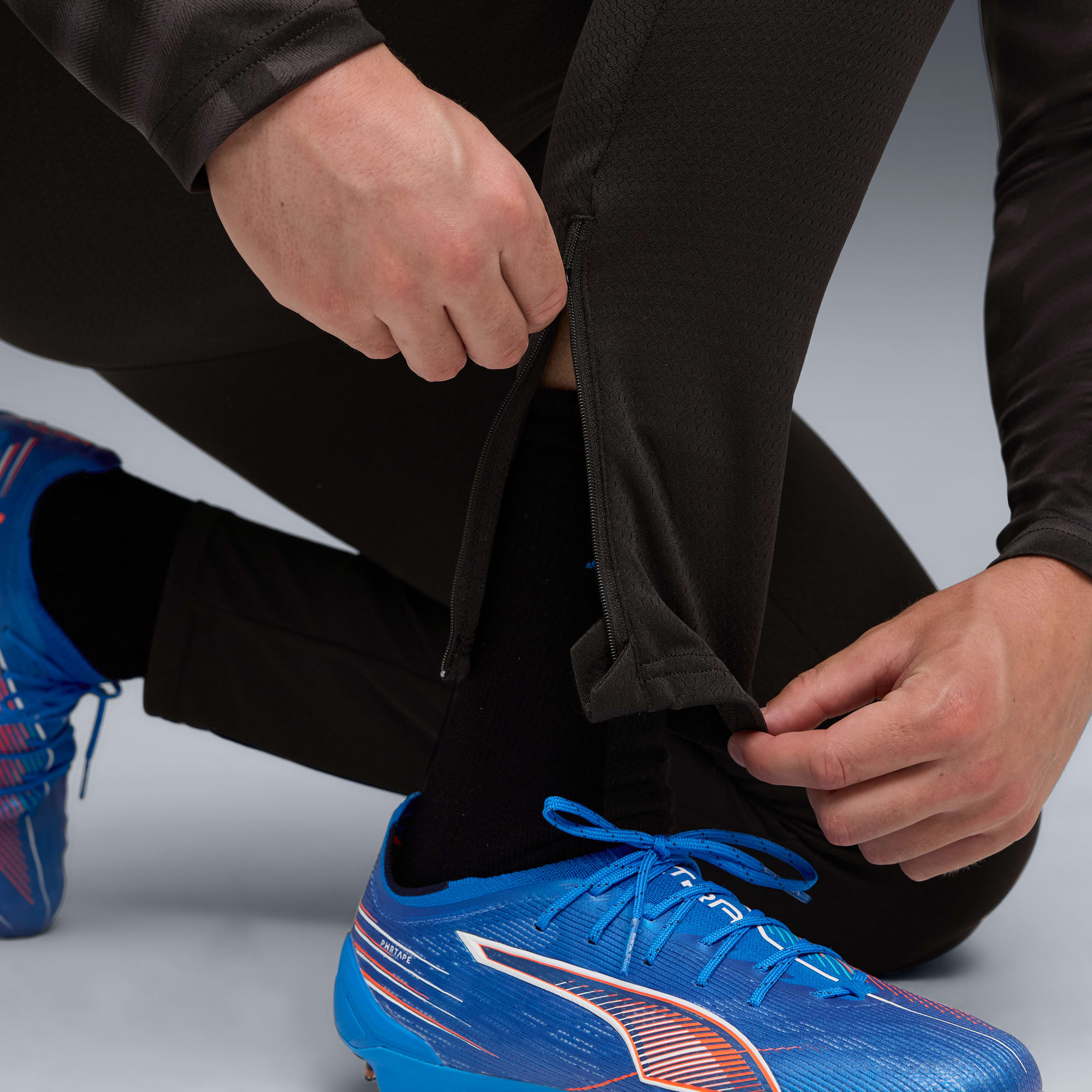 Op grijs, een persoon knielt en opent de rits van een donkere sportbroek over blauwe voetbalschoenen.