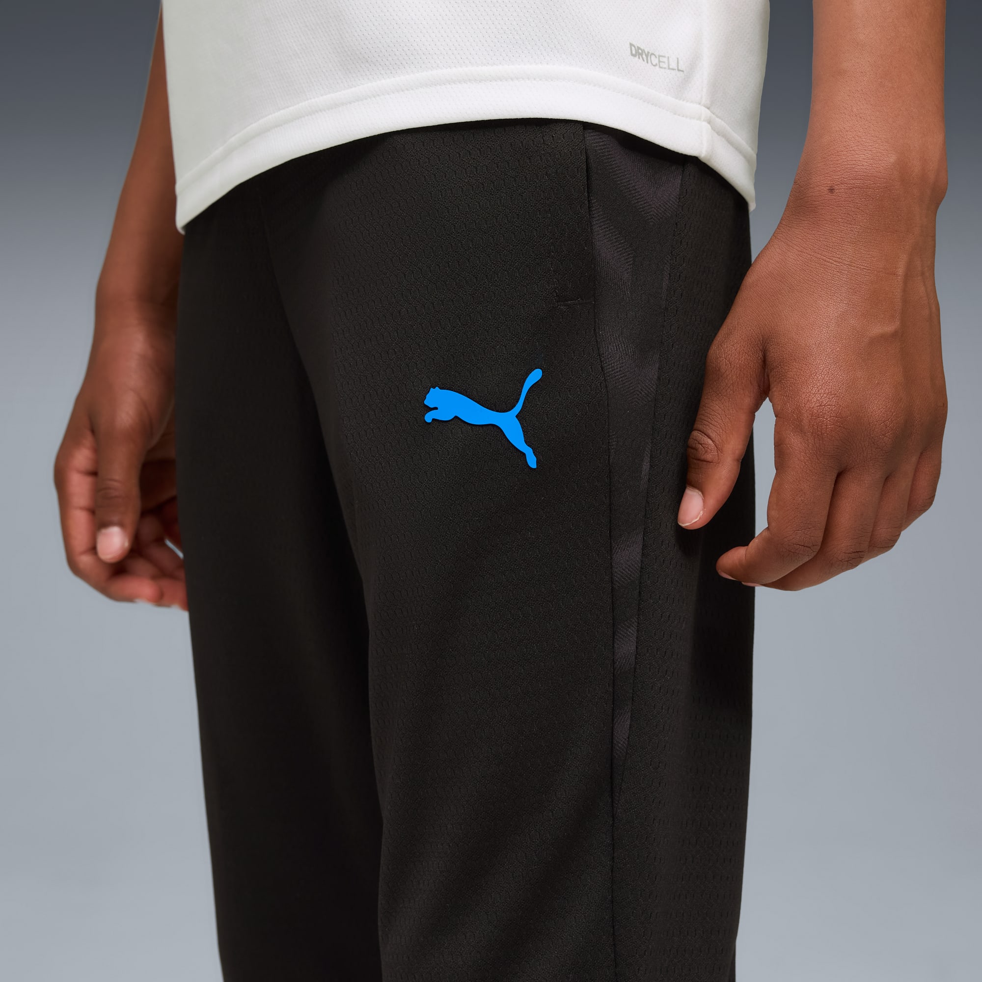Un modello indossa pantaloni da tuta neri con logo Puma blu su sfondo grigio.