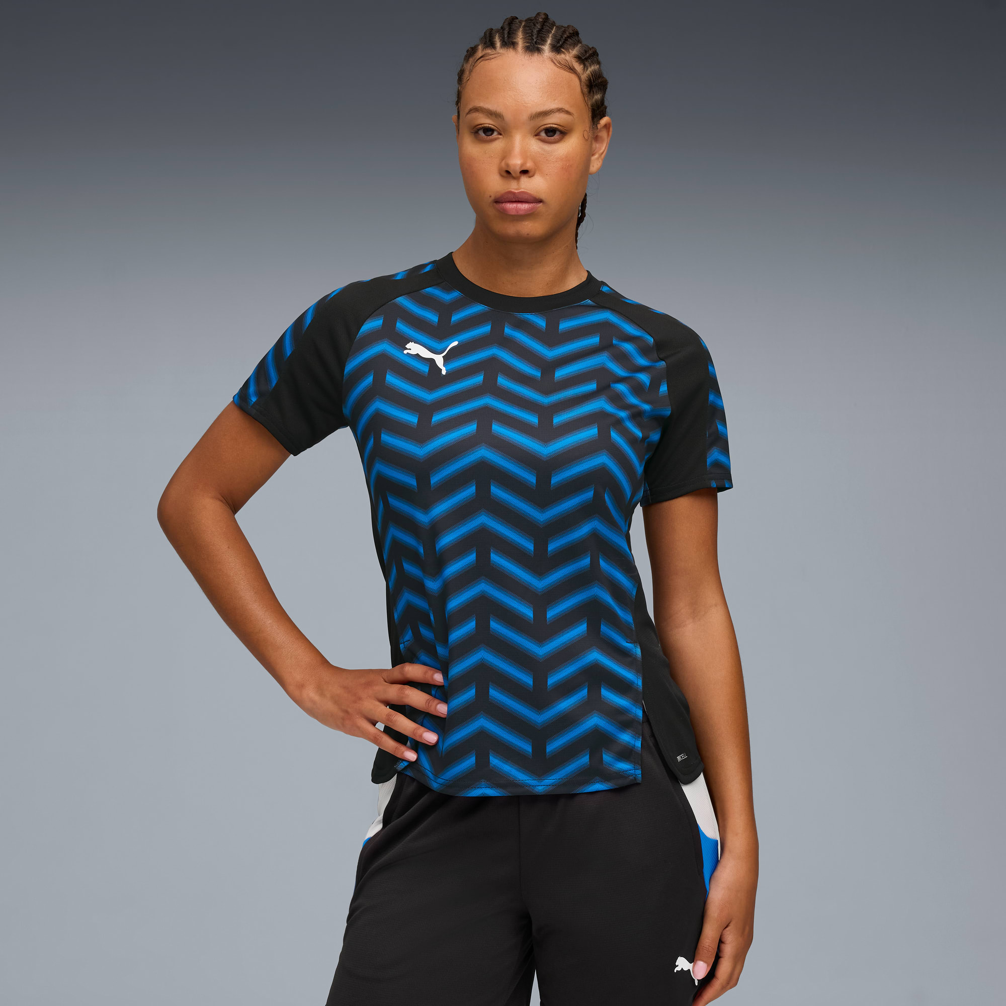 Modelka pozuje na szarym tle, ubrana w koszulkę we wzory i czarne spodenki.