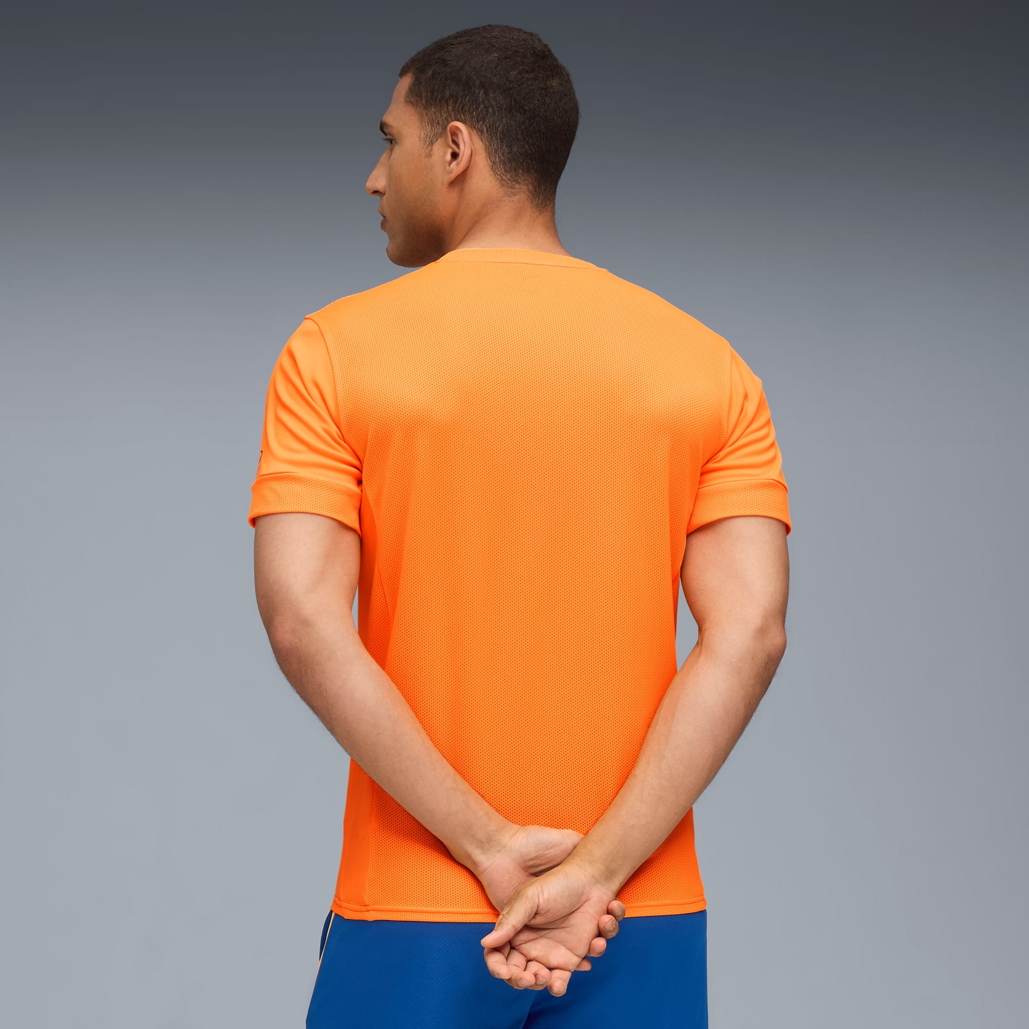 Un modello guarda a sinistra su sfondo grigio, indossa una t-shirt arancione e pantaloncini blu.