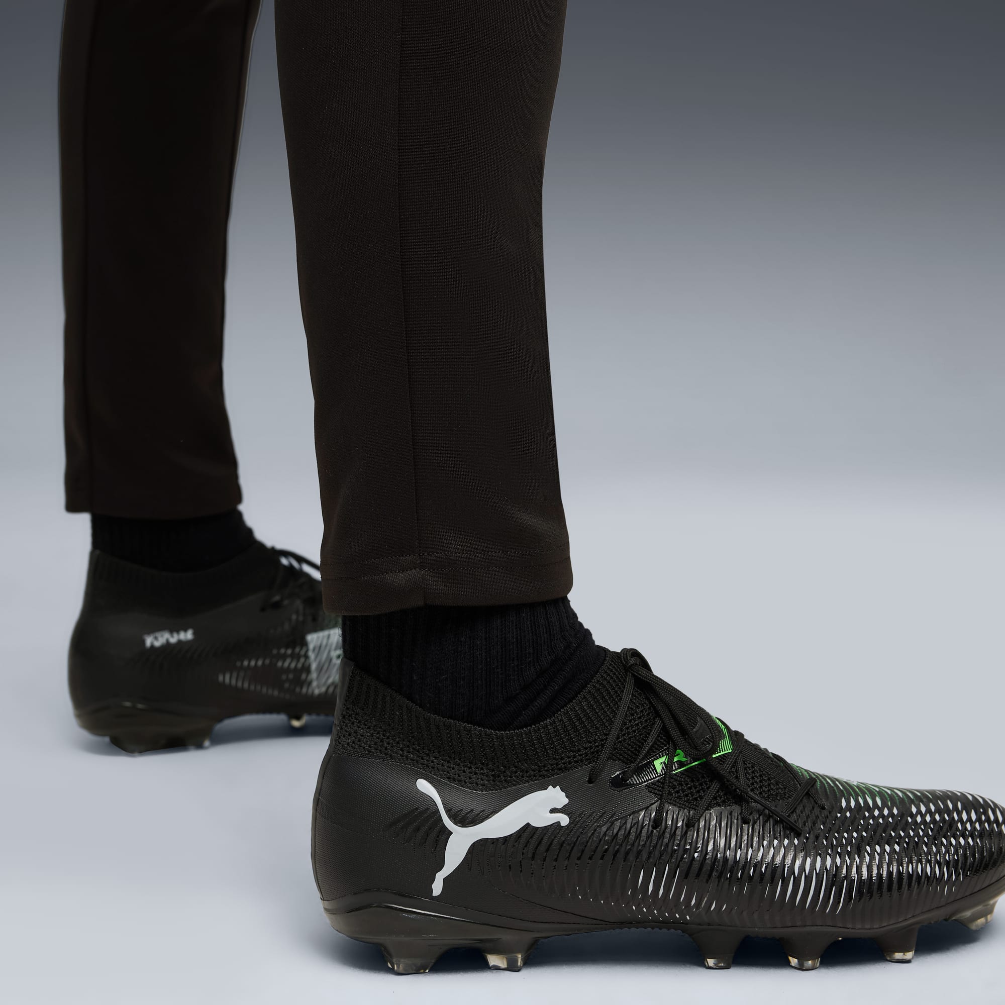 Tegen een grijze achtergrond draagt een model een zwarte broek en zwarte voetbalschoenen.
