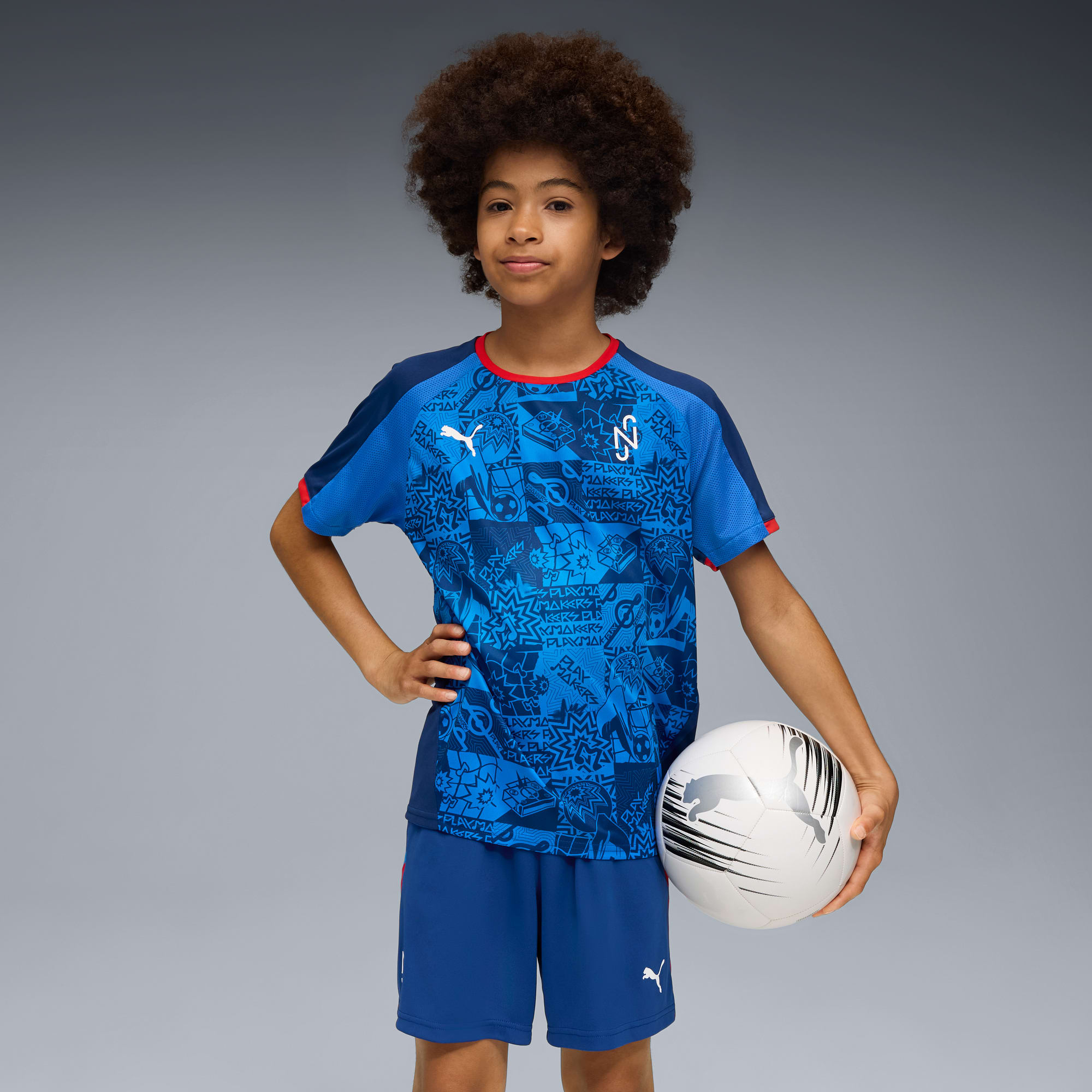 Sur fond gris, un enfant pose avec un ballon, portant un t-shirt bleu à motifs et un short.
