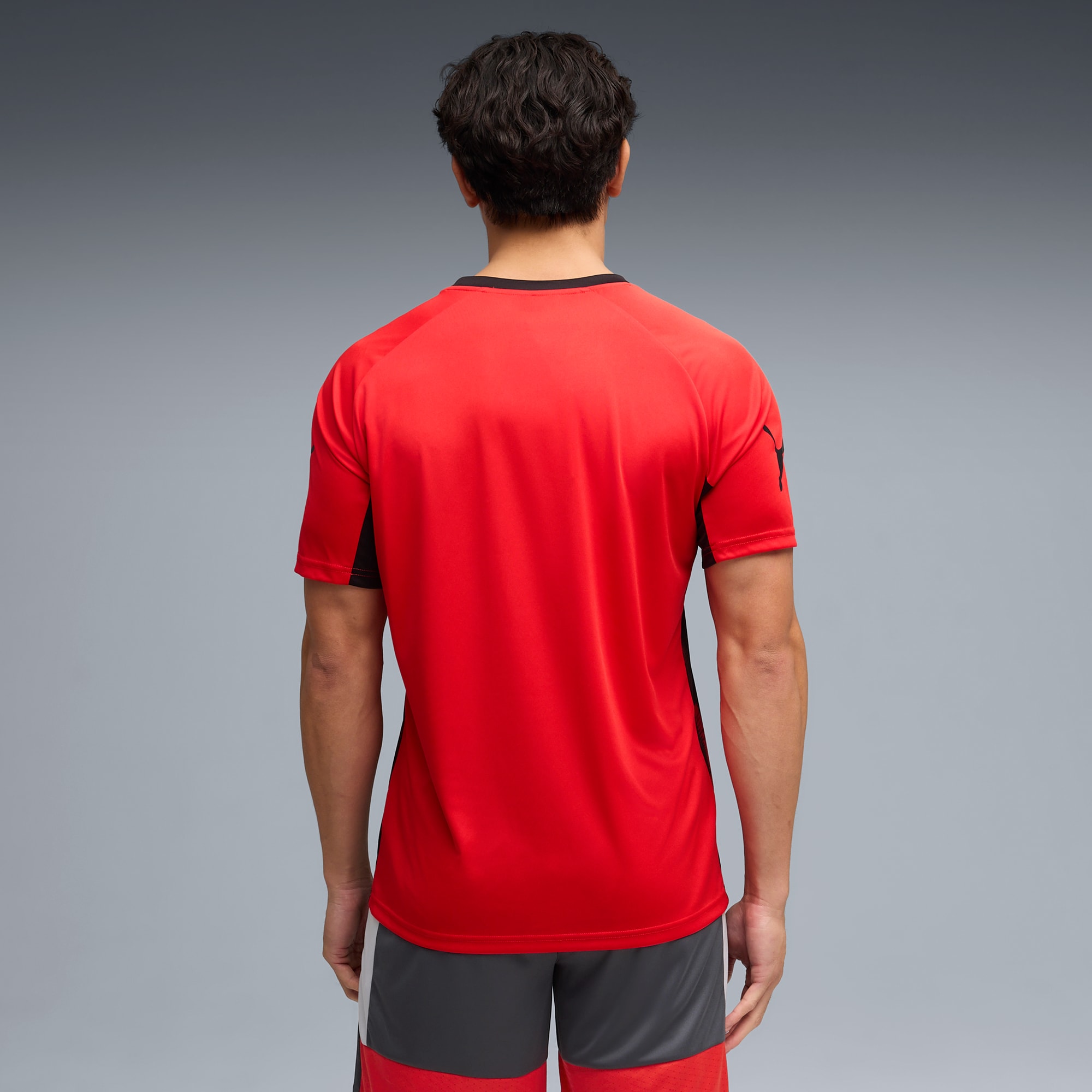 Un modelo de espaldas sobre fondo gris, con camiseta roja y pantalón corto gris.