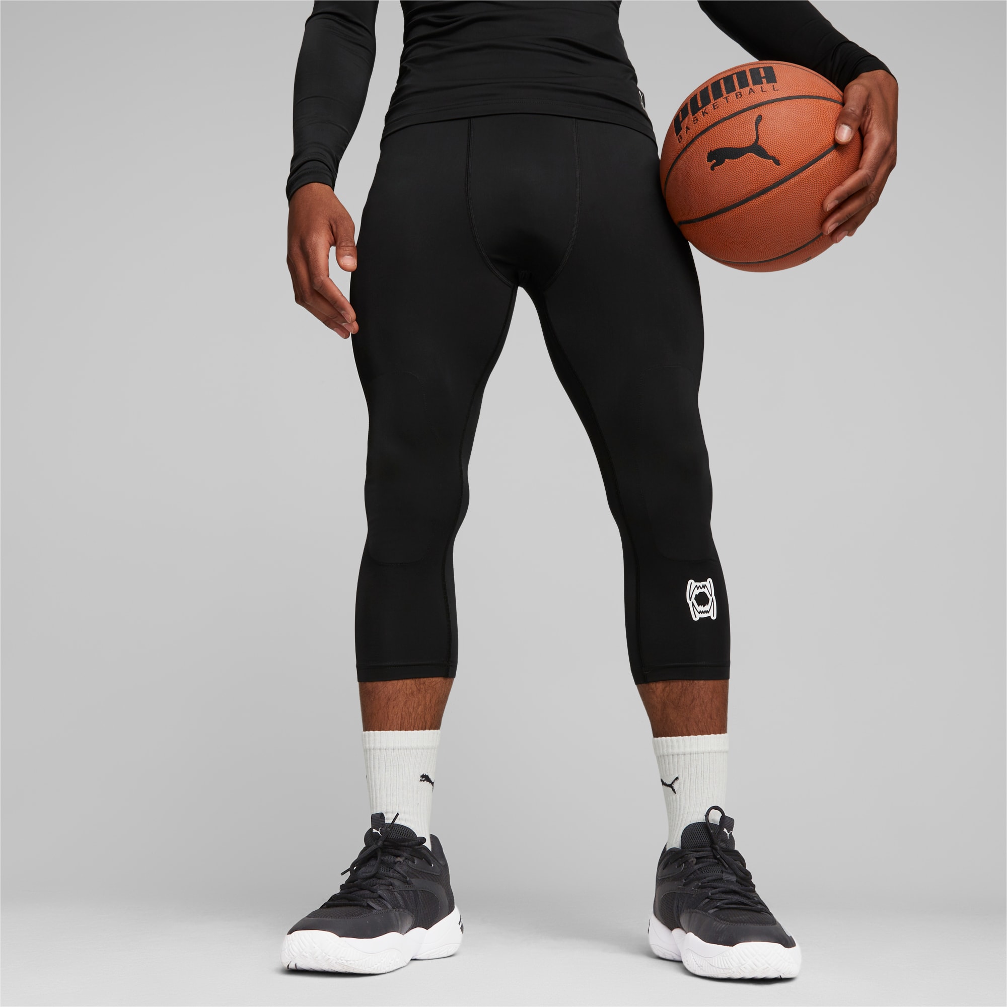 Een model houdt een basketbal vast tegen een grijze achtergrond, gekleed in zwarte sportkleding en sneakers.