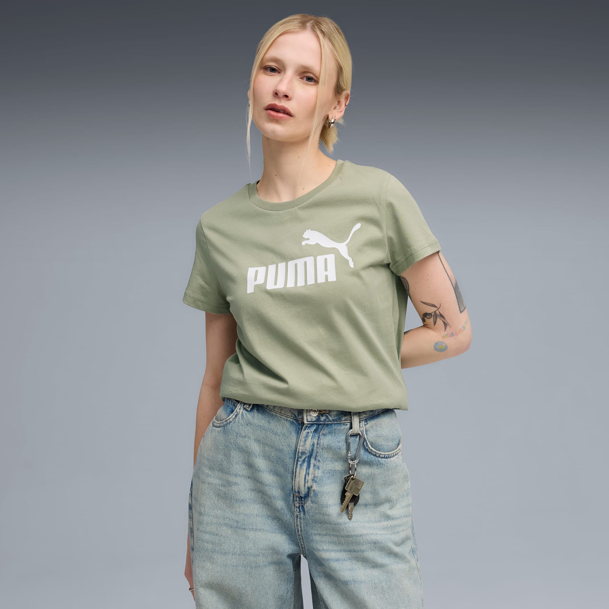 Een model poseert tegen een grijze achtergrond, gekleed in een lichtgroen logo-t-shirt en jeans.