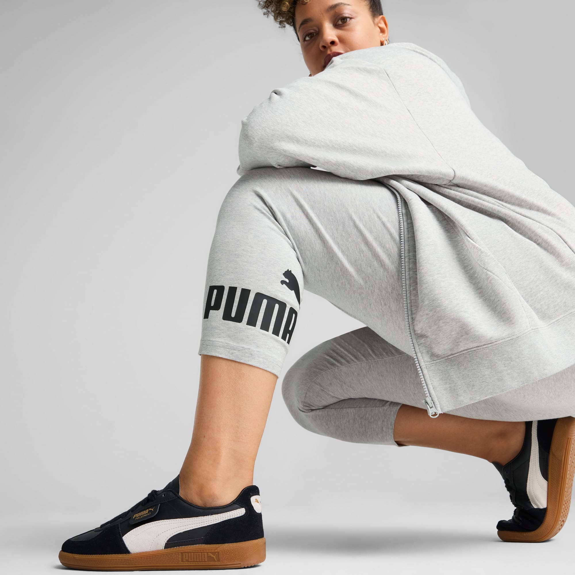 Een model hurkt voor een grijze achtergrond, gekleed in een grijs pak en zwarte hardloopschoenen.