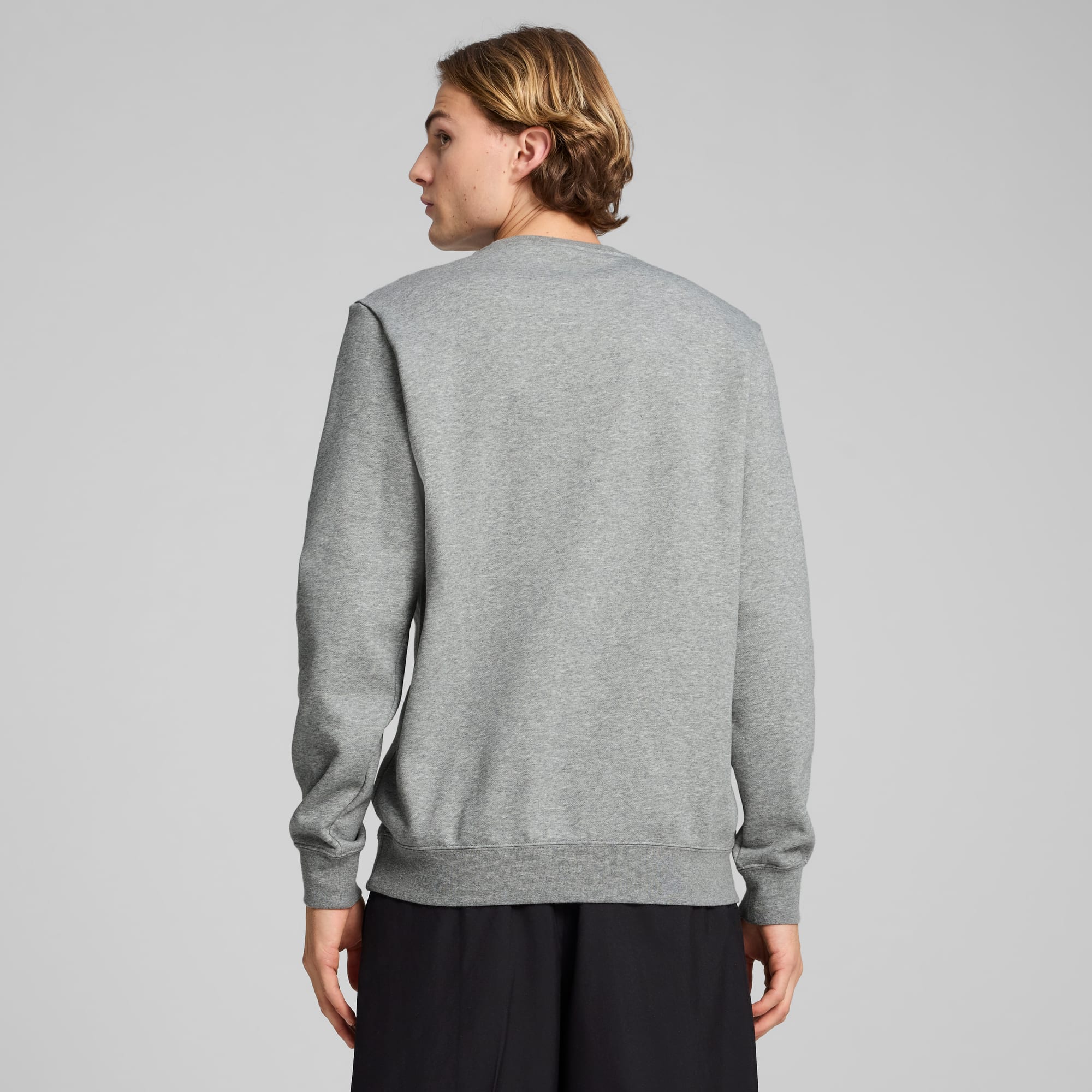 Ein Model mit grauem Sweatshirt und schwarzen Shorts vor grauem Hintergrund, Rückansicht.