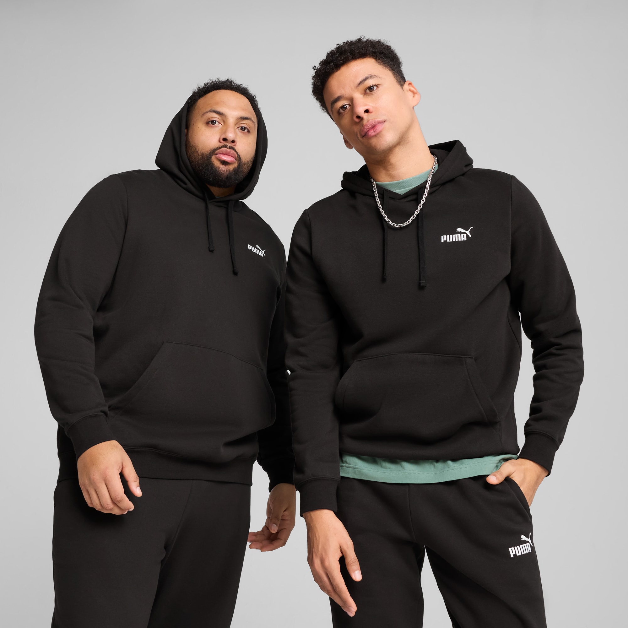 Zwei Models posieren vor grauem Hintergrund in schwarzen Hoodies und Hosen.