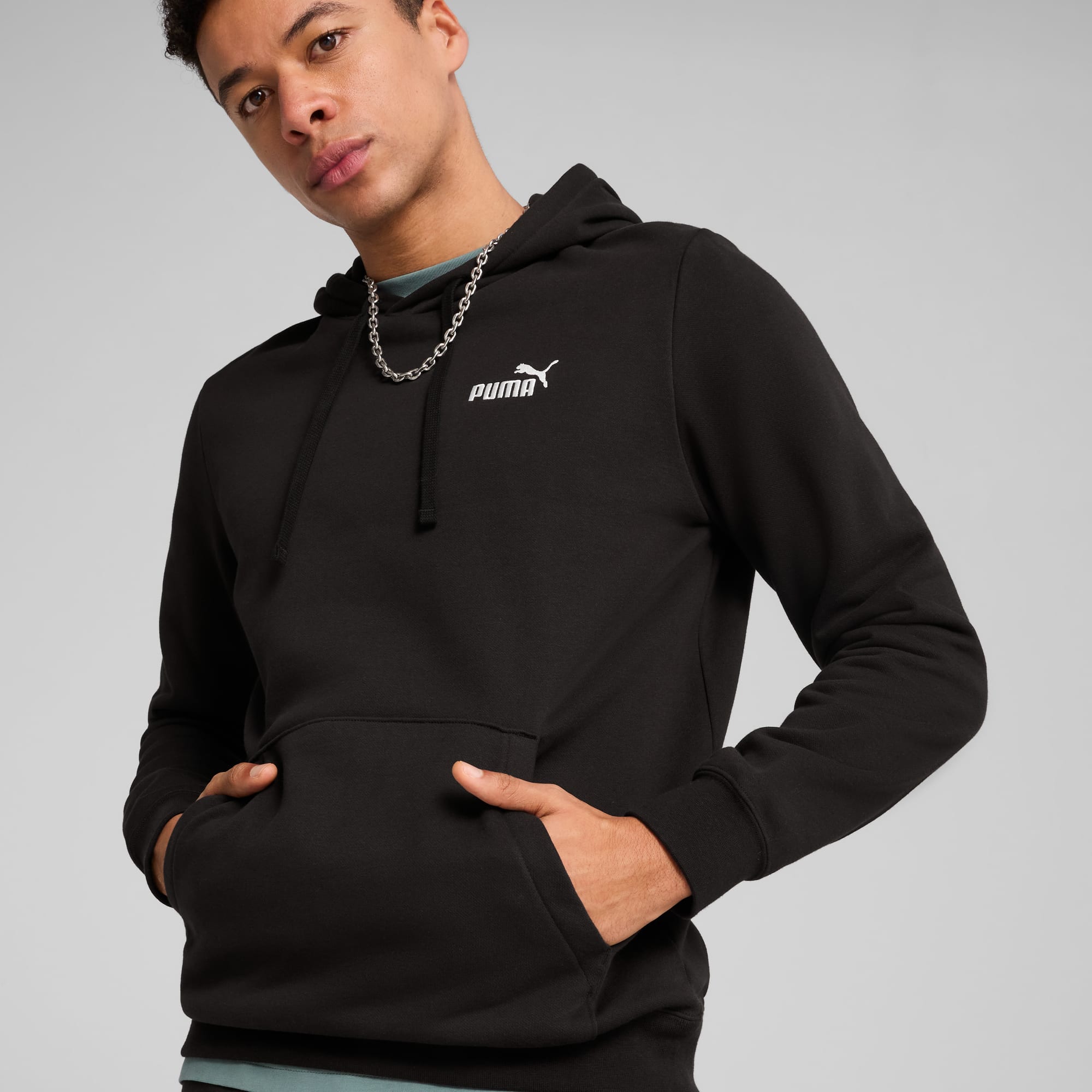 Vor grauem Hintergrund posiert ein Model mit schwarzem Hoodie und silberner Kette.