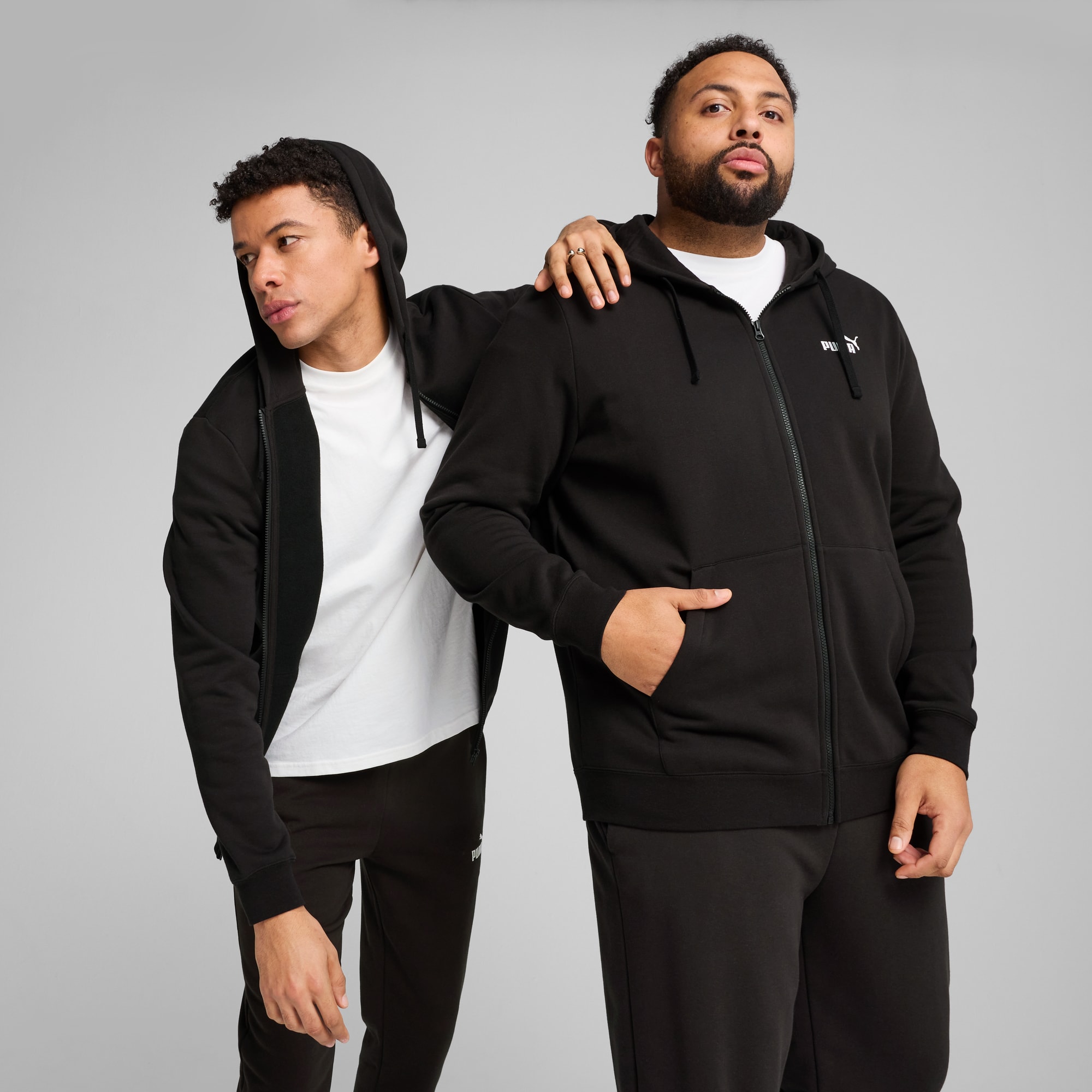 Zwei Models posieren vor grauem Hintergrund, tragen schwarze Trainingsanzüge und weiße T-Shirts.