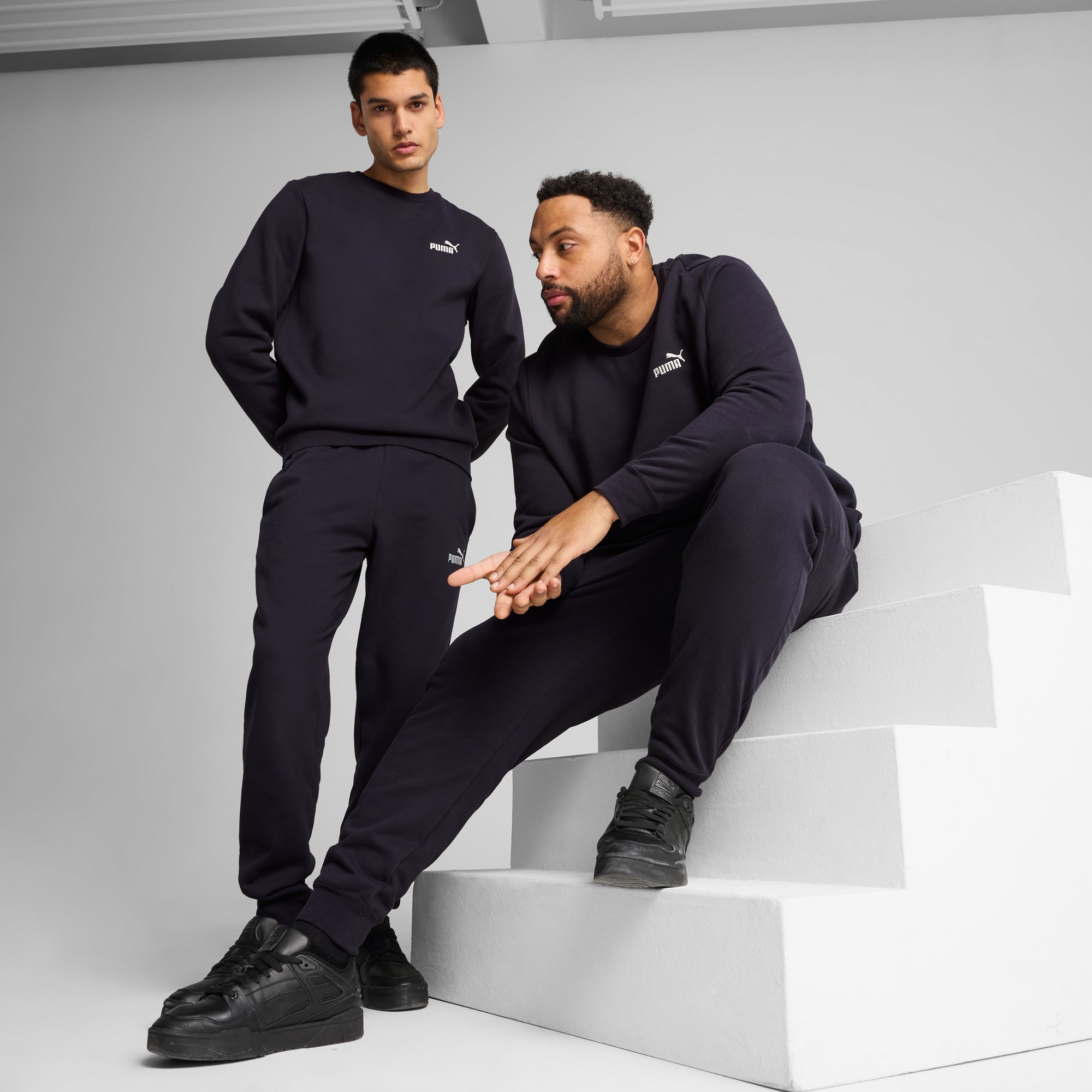 Two models pose in dark blue tracksuits and black trainers against a grey backdrop.
