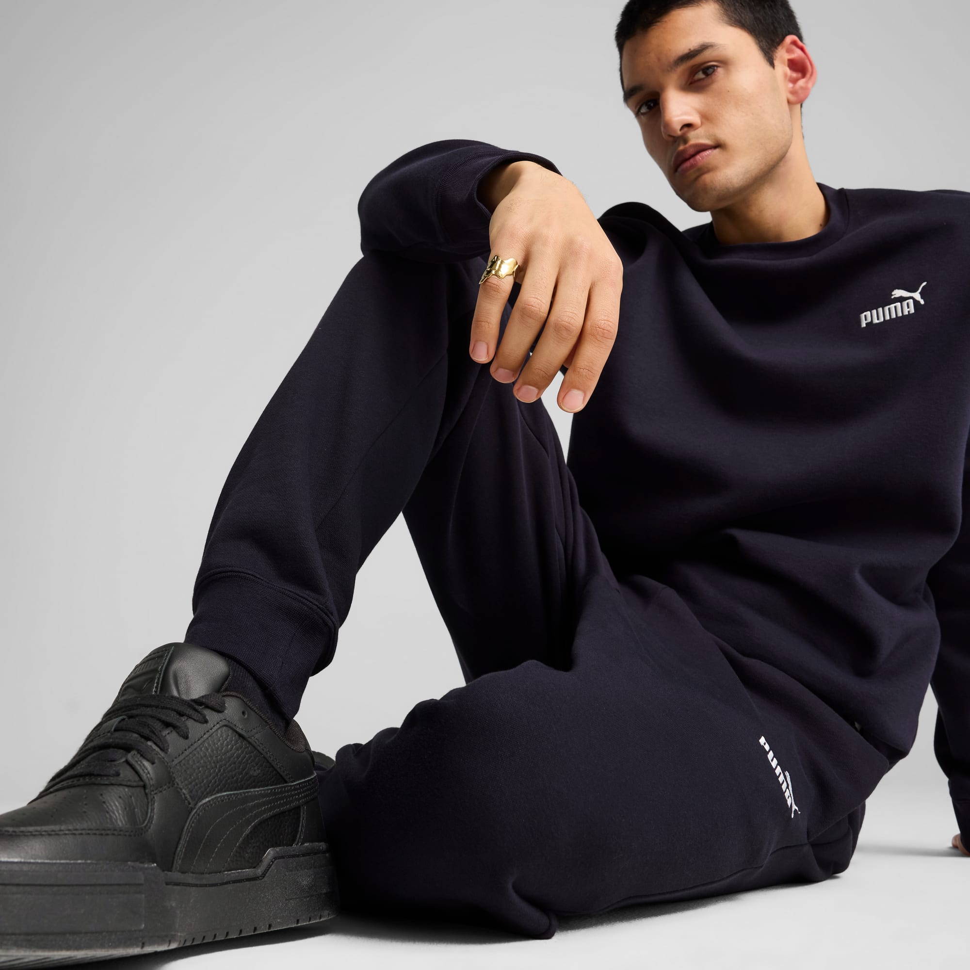 A model sits against grey, wearing a navy sweatshirt, joggers, black trainers and a ring.