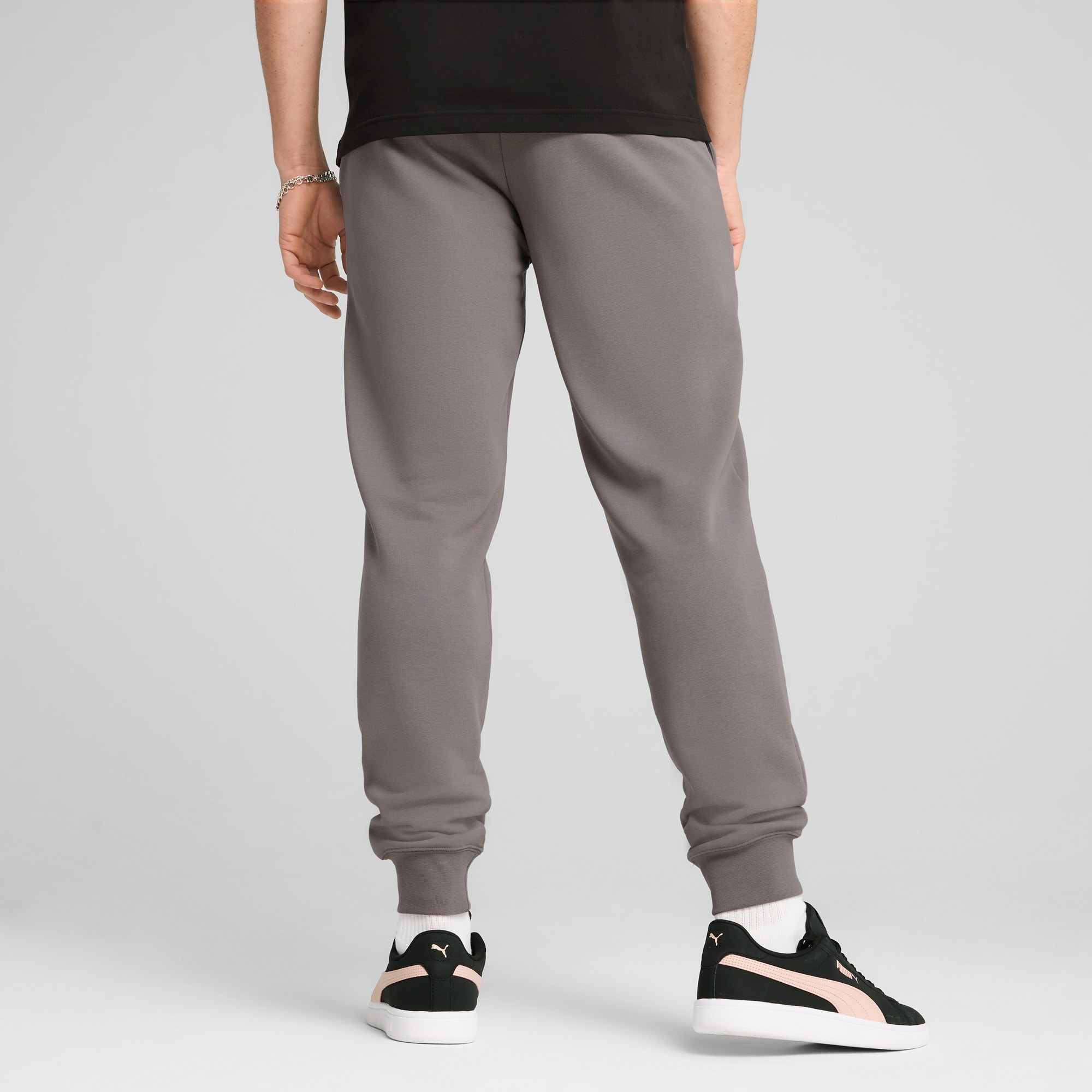 Een model staat met de rug naar de camera op een grijze achtergrond, met grijze broek en zwarte sneakers.