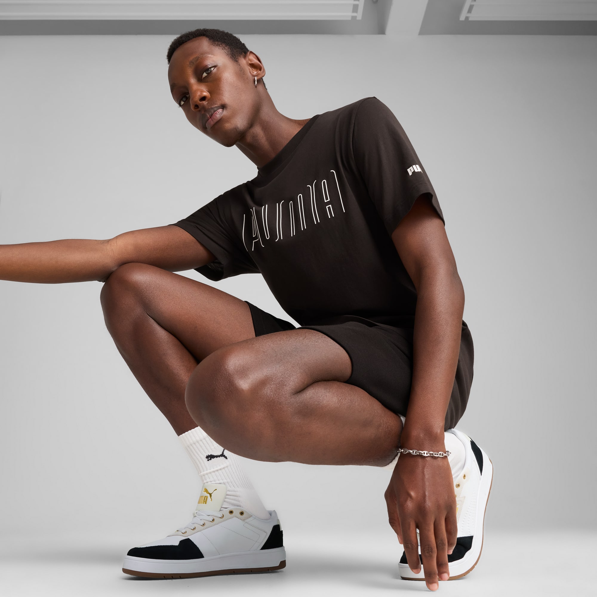 Su sfondo grigio, un modello accovacciato indossa t-shirt, shorts e scarpe da ginnastica.