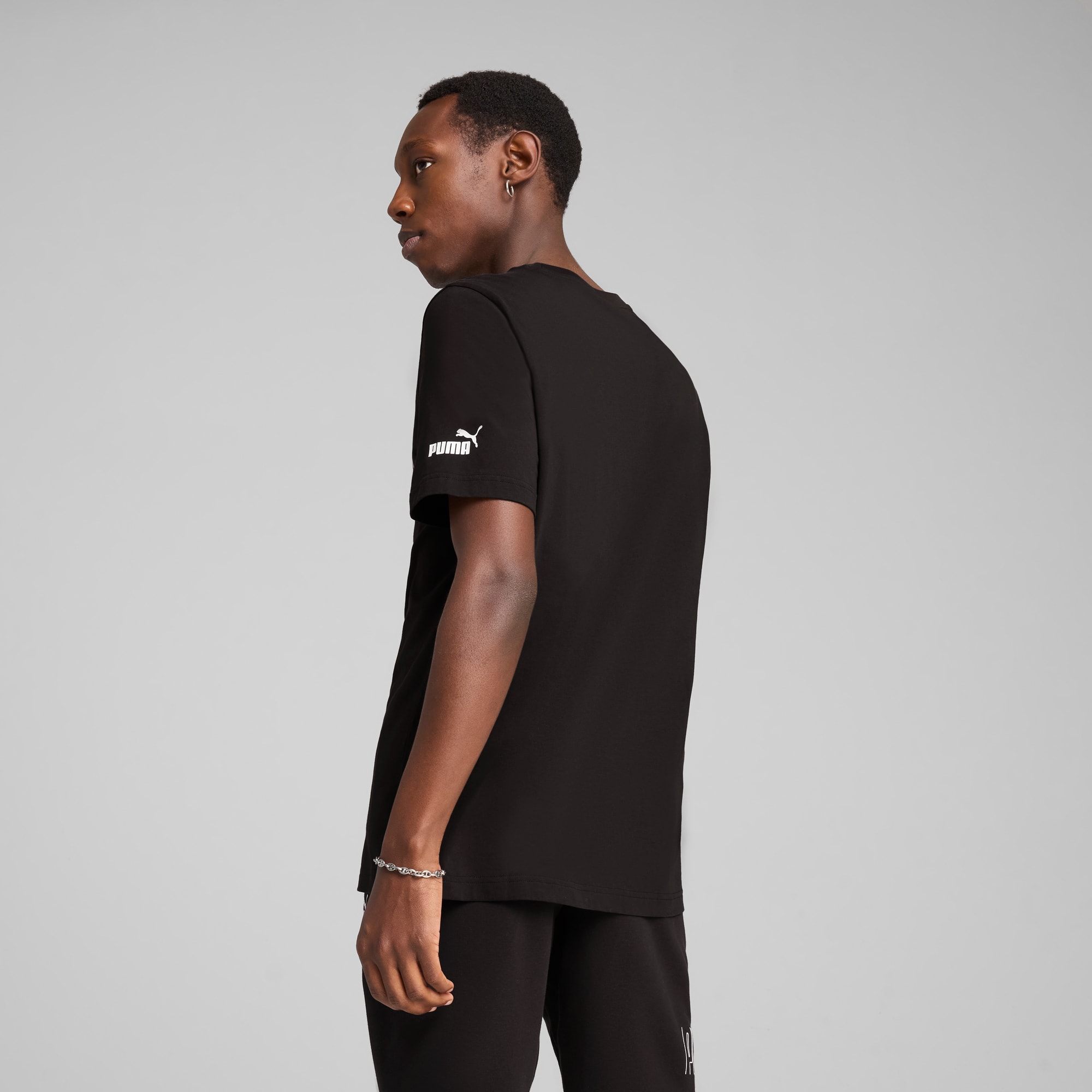 Un modello su sfondo grigio indossa una t-shirt nera e pantaloni neri, guardando di lato.