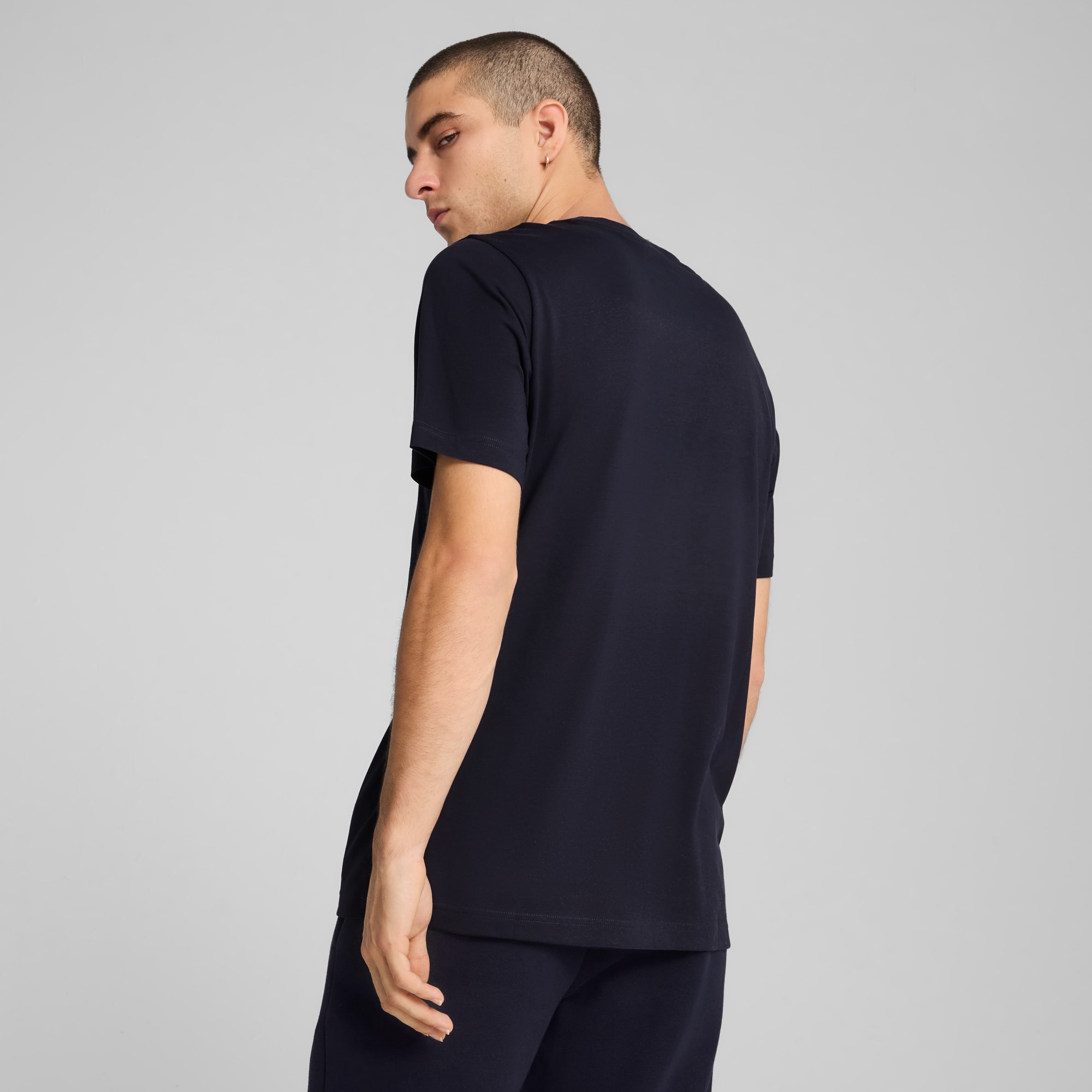 Un modèle pose sur fond gris, portant un t-shirt et un pantalon sombres, regardant par-dessus son épaule.