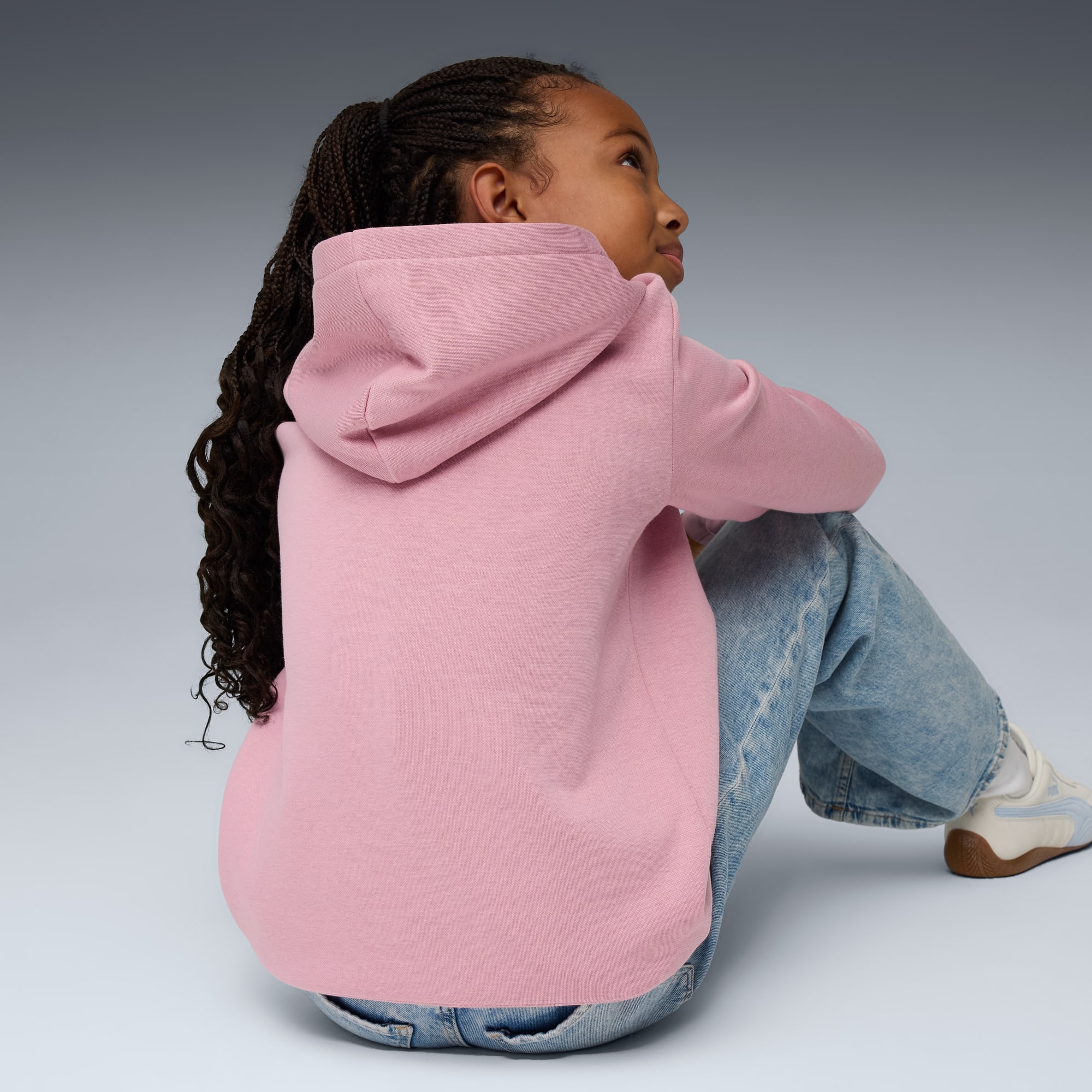 Su sfondo grigio, una modella siede indossando una felpa rosa con cappuccio e jeans, guardando verso l'alto.