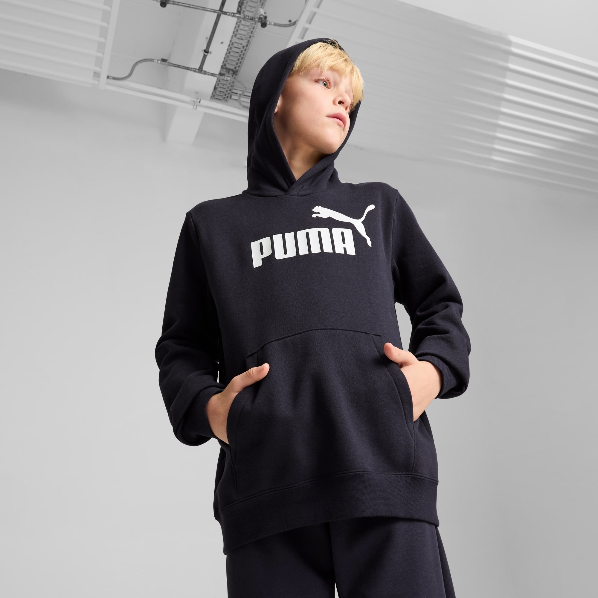 Ein Model posiert vor grauem Hintergrund und trägt ein dunkles Kapuzenshirt mit weißem Puma-Logo.