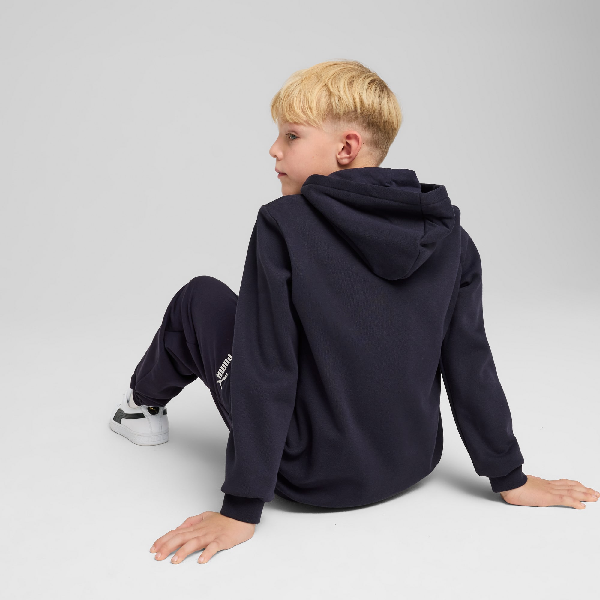 Auf Grau sitzt ein Kind mit dunklem Kapuzenpullover, passender Hose und weißen Turnschuhen.